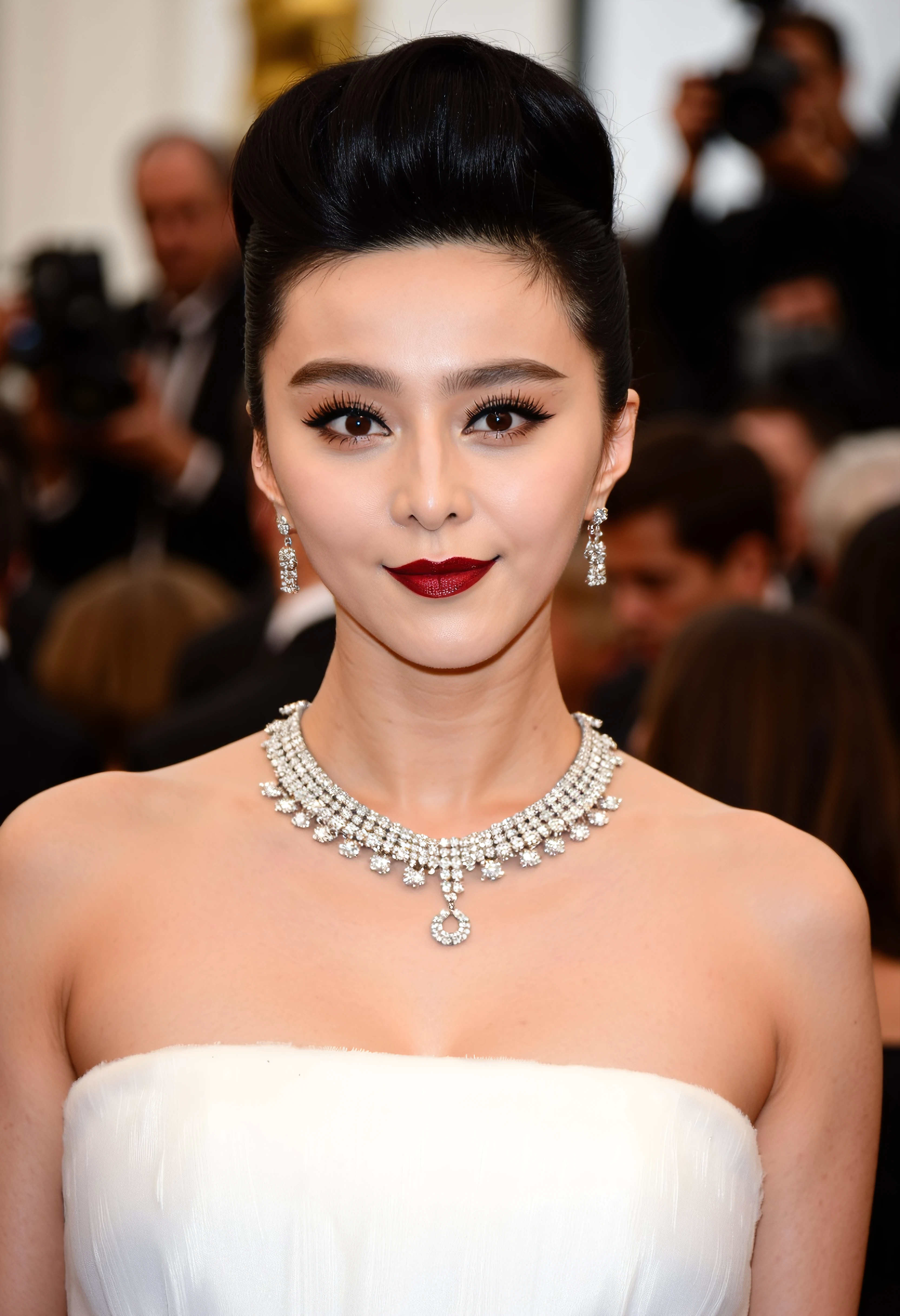 The image is a close-up portrait of a young woman at an event. She is wearing a white strapless dress with a statement necklace and earrings. Her hair is styled in a sleek updo with dark hair framing her face. She has a bold red lipstick on and her makeup is done in a dramatic style with dark eyeliner and winged eyeliner. The background is blurred but it appears to be a crowd of people at a formal event. The woman is looking directly at the camera with a serious expression.