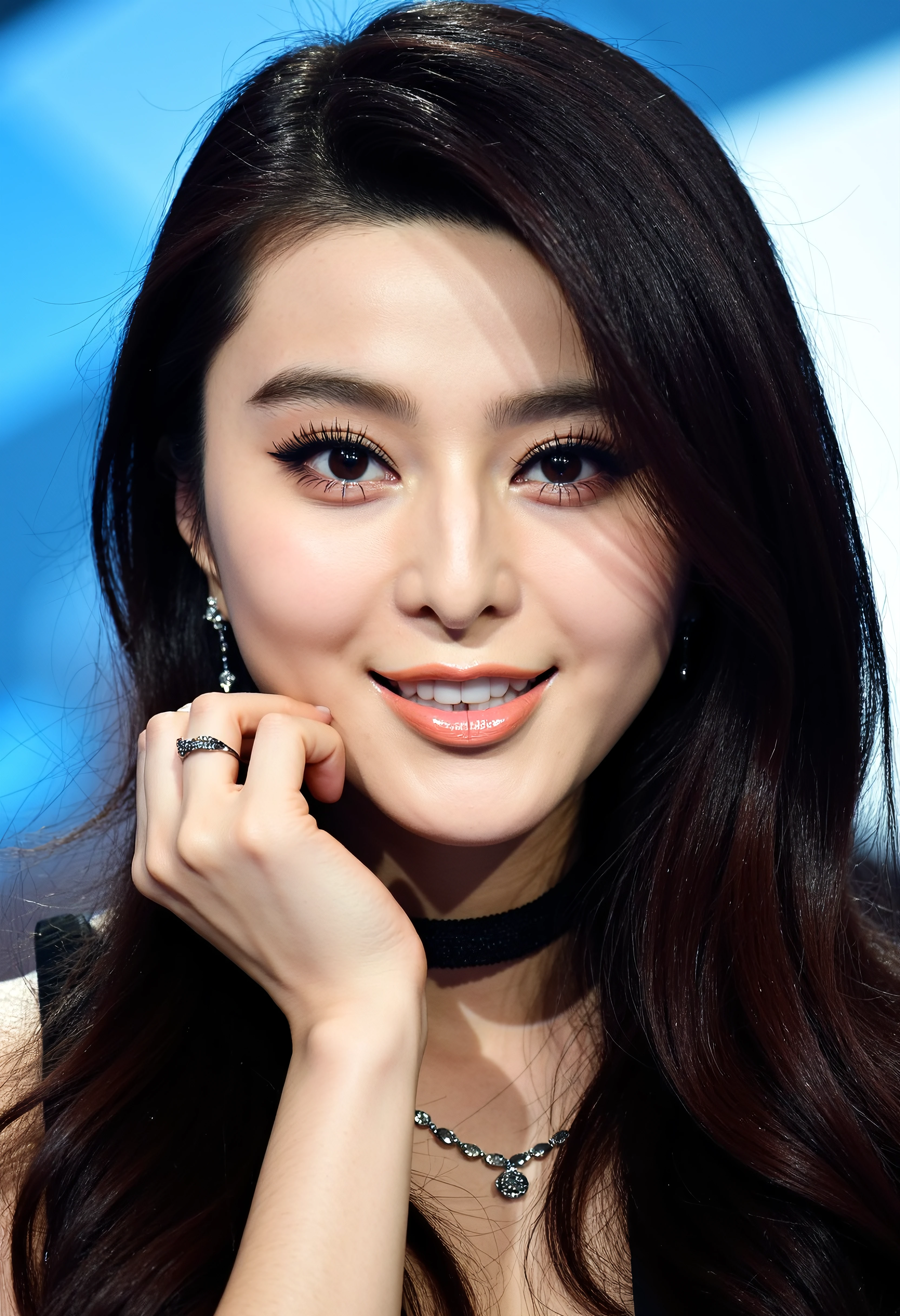 The image is a close-up portrait of a young woman with long dark hair. She is smiling and looking directly at the camera. Her hair is styled in loose waves and falls over her shoulders. She has a ring on her finger and is wearing a black choker necklace. The background is blurred but it appears to be an indoor setting with a blue and white color scheme. The woman's makeup is natural-looking and she has a subtle smoky eye makeup look. She appears to have a slight smile on her lips.