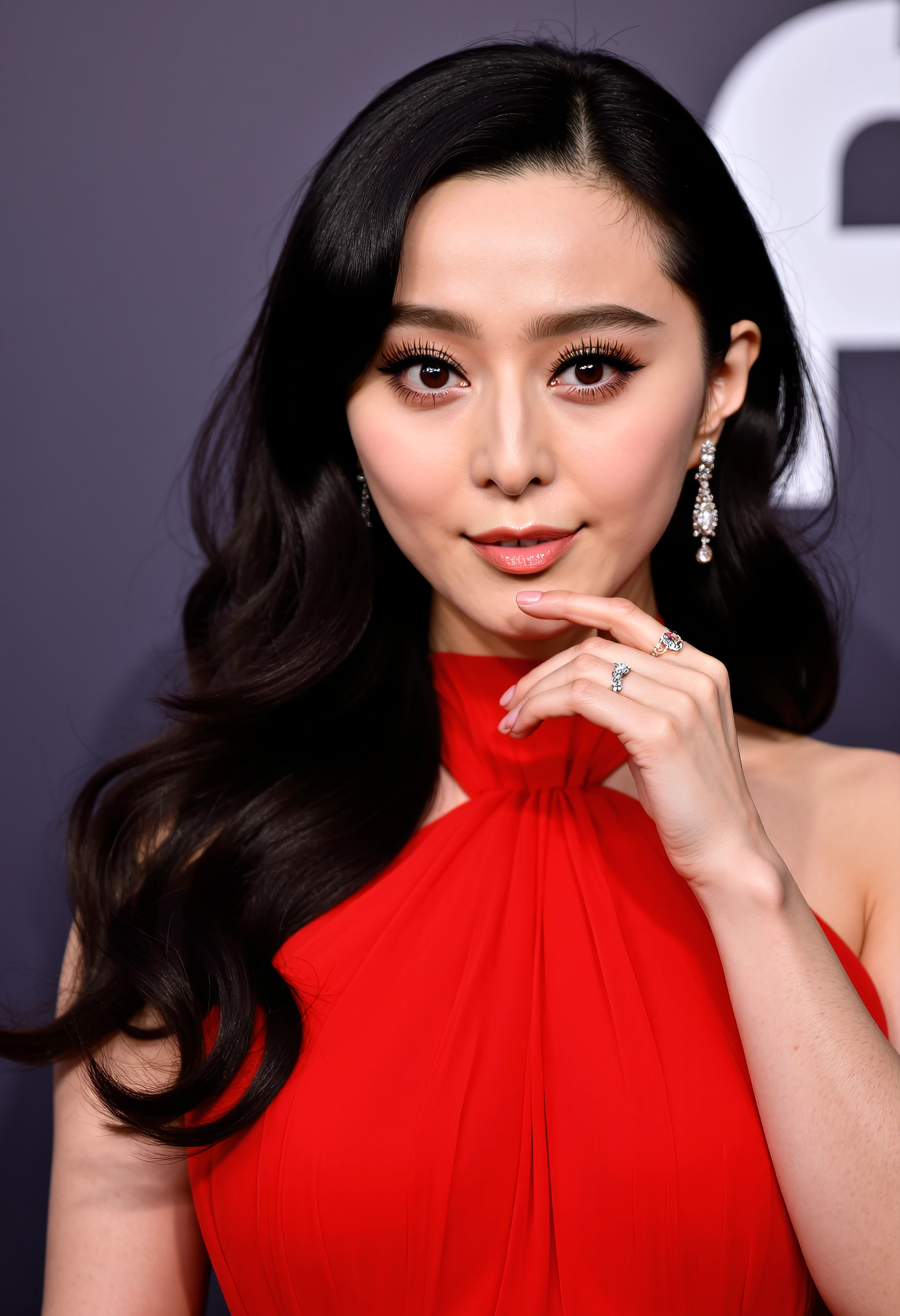 The image is a portrait of a young woman with long dark hair. She is wearing a red dress with a high neckline and long sleeves. Her hair is styled in loose waves and falls over her shoulders. She has a serious expression on her face and is looking directly at the camera with a slight smile. Her makeup is subtle and natural-looking with a hint of pink blush on her cheeks and a nude lip. Her eyes are accentuated with a smoky eye makeup look and her eyebrows are groomed and filled in. She also has a pair of statement earrings and a ring on her finger. The background is