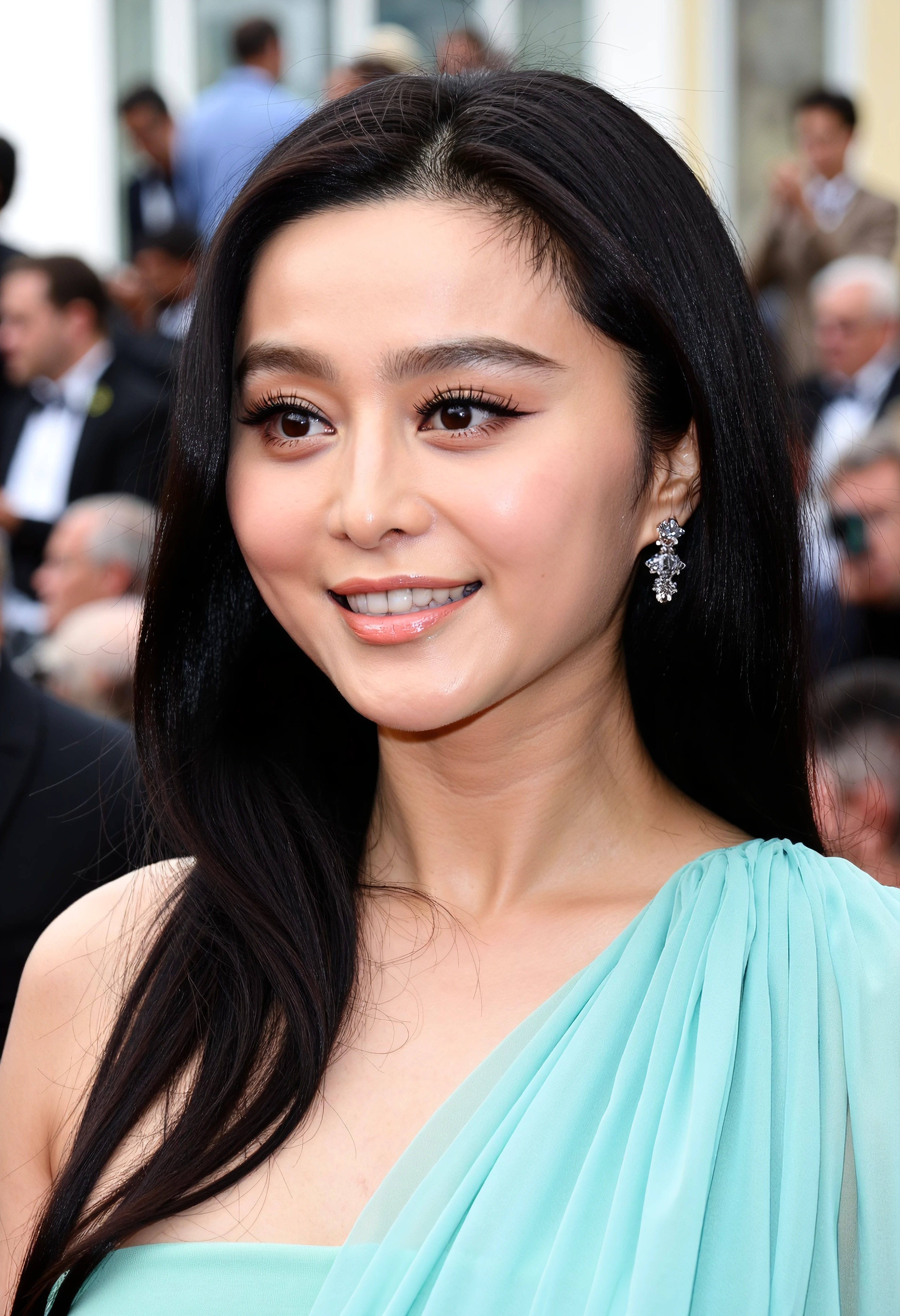 The image is a close-up portrait of a young woman with long dark hair. She is smiling and looking directly at the camera. Her hair is styled in loose waves and falls over her shoulders. She has a subtle smokey eye makeup look with dark eyeliner and a nude lip color. Her makeup is natural-looking with a hint of pink blush on her cheeks and a subtle smoky eye. She also has a pair of dangling earrings on her left ear. The background is blurred but it appears to be an outdoor event with people and buildings visible. The woman is wearing a light blue dress with a draped neck