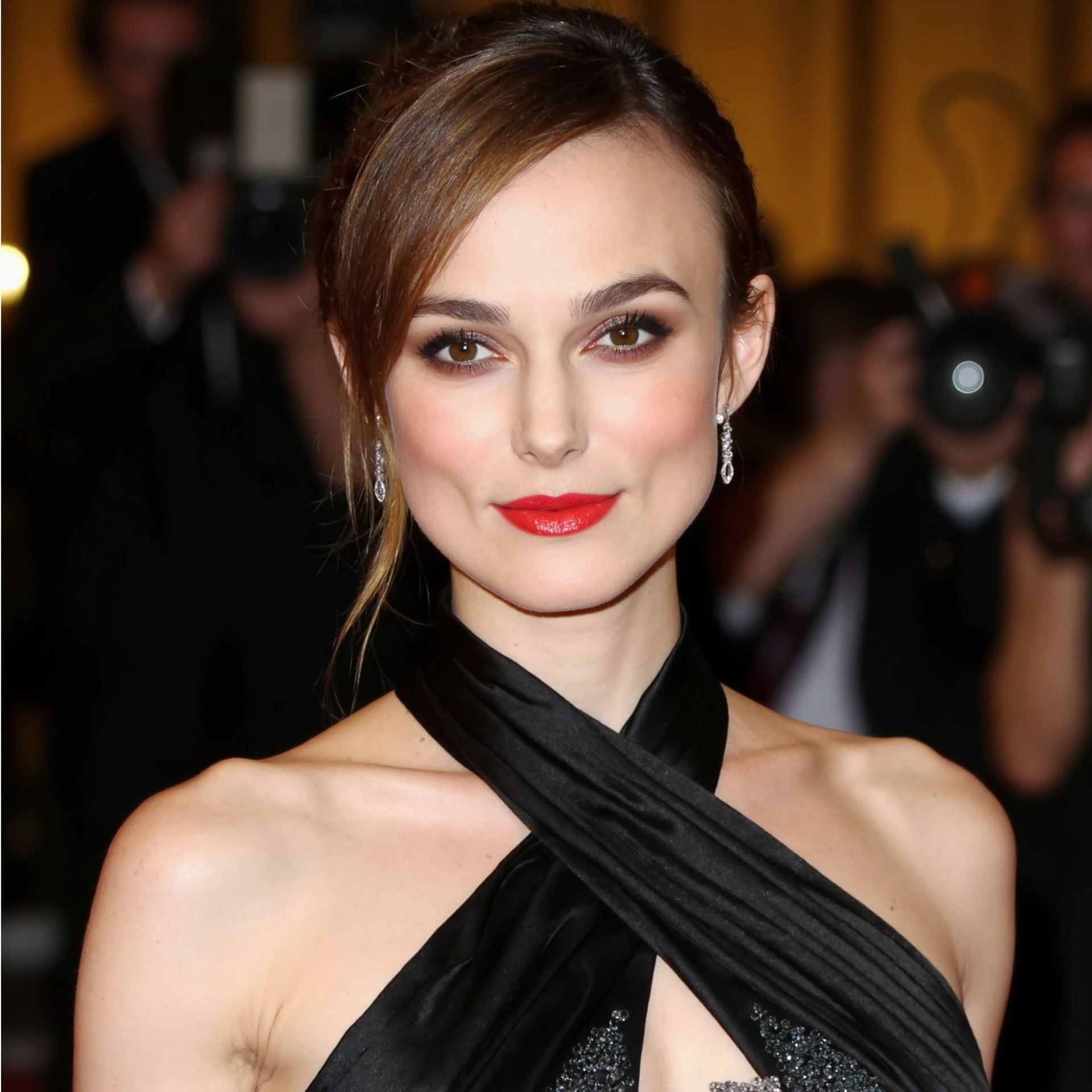 A close-up portrait of  keira knightley a brunette woman at a red carpet event, wearing an elegant closed neckline black dress. The dress features intricate silk detailing along the neckline and a subtle shimmer that catches the light. Her makeup is flawlessly applied, with a classic red lip and a smoky eye, enhancing her natural beauty. Her hair falling down besides her face in a very stylish way, with a few loose strands framing her face. She is wearing elegant diamond earrings that sparkle against her skin. The background is softly blurred, with hints of flashing cameras and glamorous lighting, drawing all attention to her poised and radiant expression. The overall mood of the portrait is one of sophistication and timeless elegance <lora:keira_knightley_r8_1600_flux_lora_v1_000002600:1>