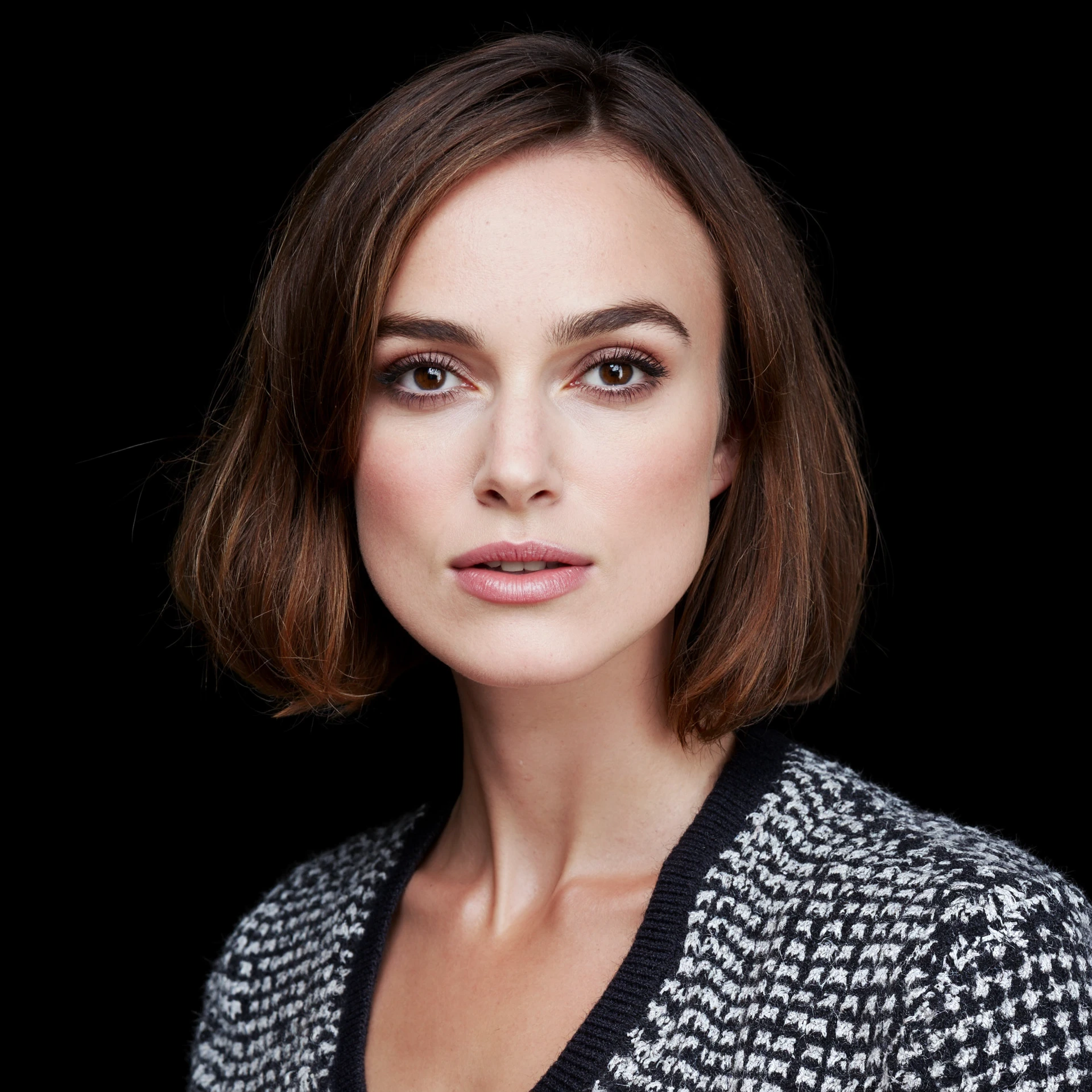 A closeup portrait photo of a stunning woman  keira knightley with medium length hair who gazing directly at the camera with a neutral expression.She is wearing a multicolored knitted sweater, against a dark background.Spotlight illumination. She is wearing clear lipgloss. <lora:keira_knightley_r8_1600_flux_lora_v1:1>