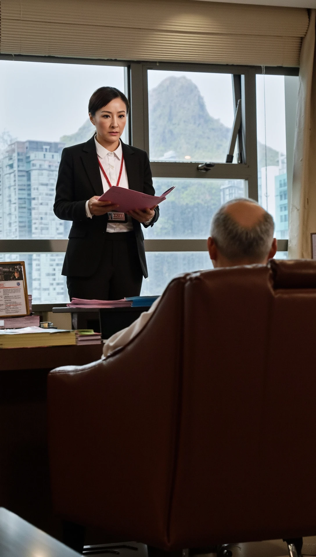 A determined Hong Kong ICAC investigator standing firmly in the office of a high-ranking government official, questioning him about corruption. 

The luxurious office is adorned with expensive art, leather furniture, and large windows showcasing the city skyline. 

The corrupt official, visibly panicked, 60 years old sits behind a grand wooden desk, while the investigator, dressed in a professional suit, holds a folder of incriminating evidence, their expression stern and unwavering.

The investigator is an old woman in a black suit and white shirt.

She has a red lanyard around her neck with a name tag attached to it.