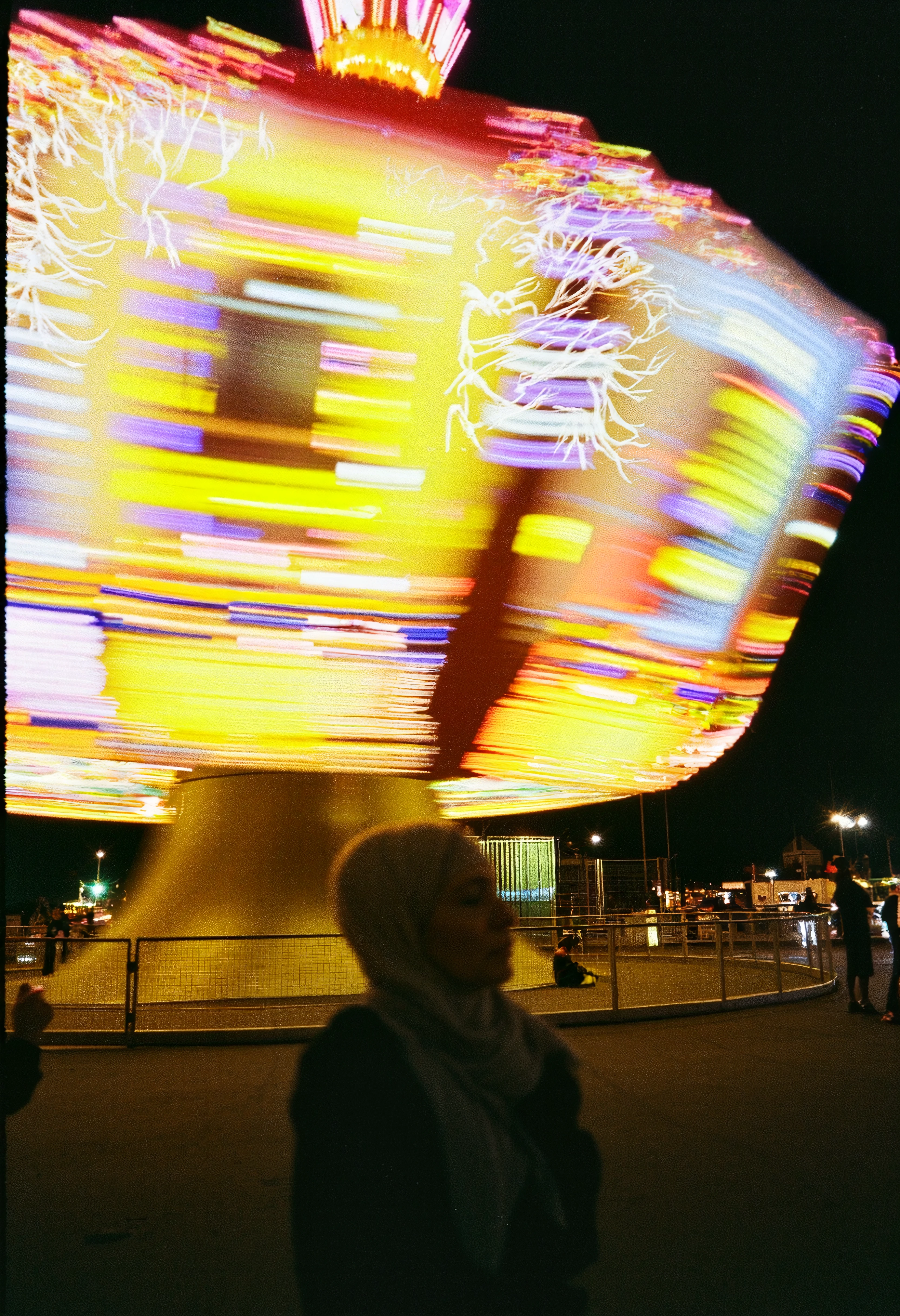 A carnival ride in motion, captured with colorful lights swirling and blurring as it spins. The motion blur of the ride creates an abstract, energetic feeling, while the vibrant colors of yellow, red, and blue streak across the frame. The chaotic energy of the scene contrasts with the stillness of the people watching below. Herbst photo, candid, color contrast, motion blur, analog texture, film grain.