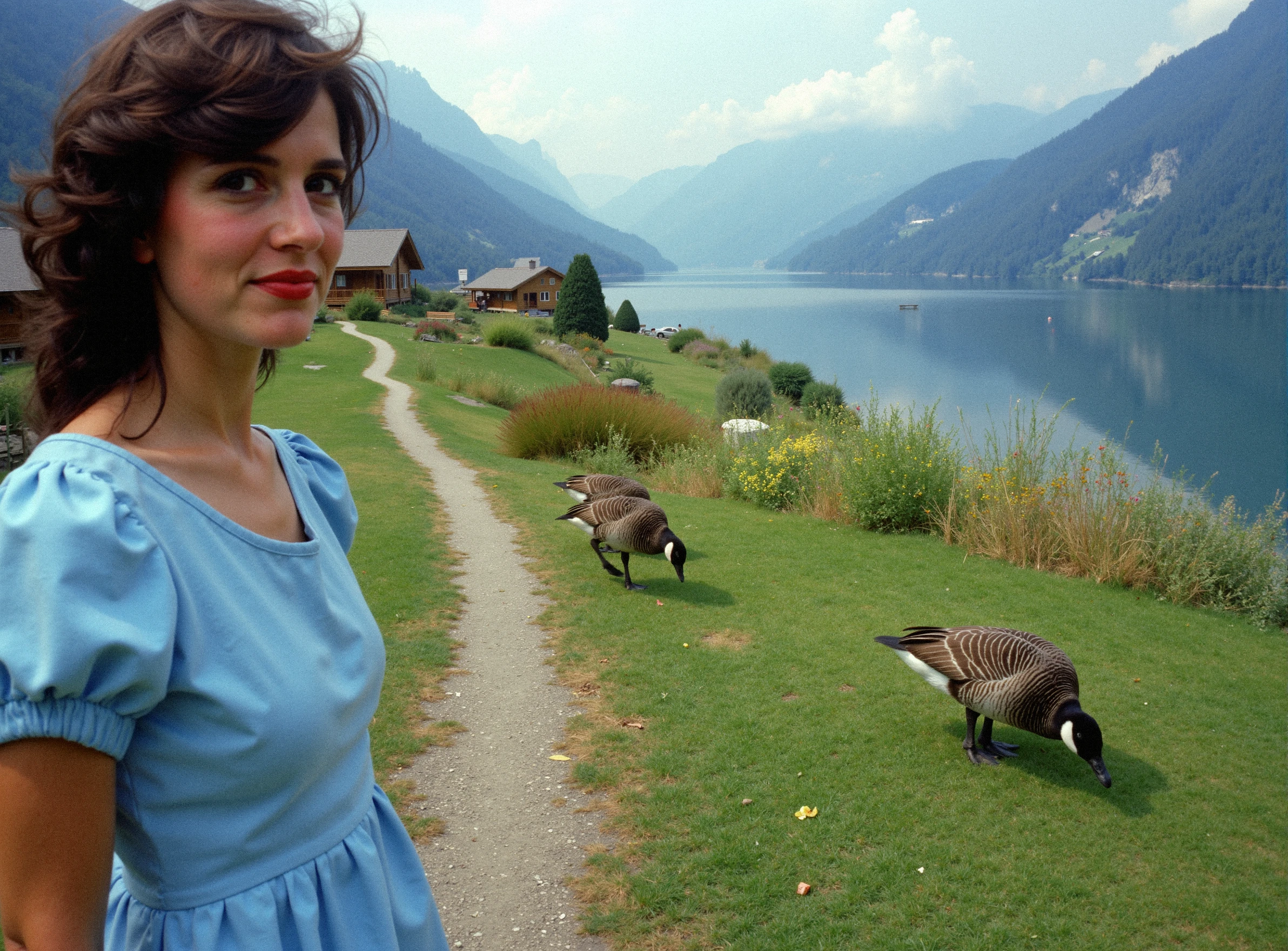 3ighti3s, closeup on a woman's face in focus, she's wearing a blue dress on a valley trail, facing the camera, shot on Kodachrome film, using a 35mm sharp lens, in the background view there are cabins and a big lake with geese. Some of the geese are walking on the grass around bushes of indigenous flowers, swiss alps.