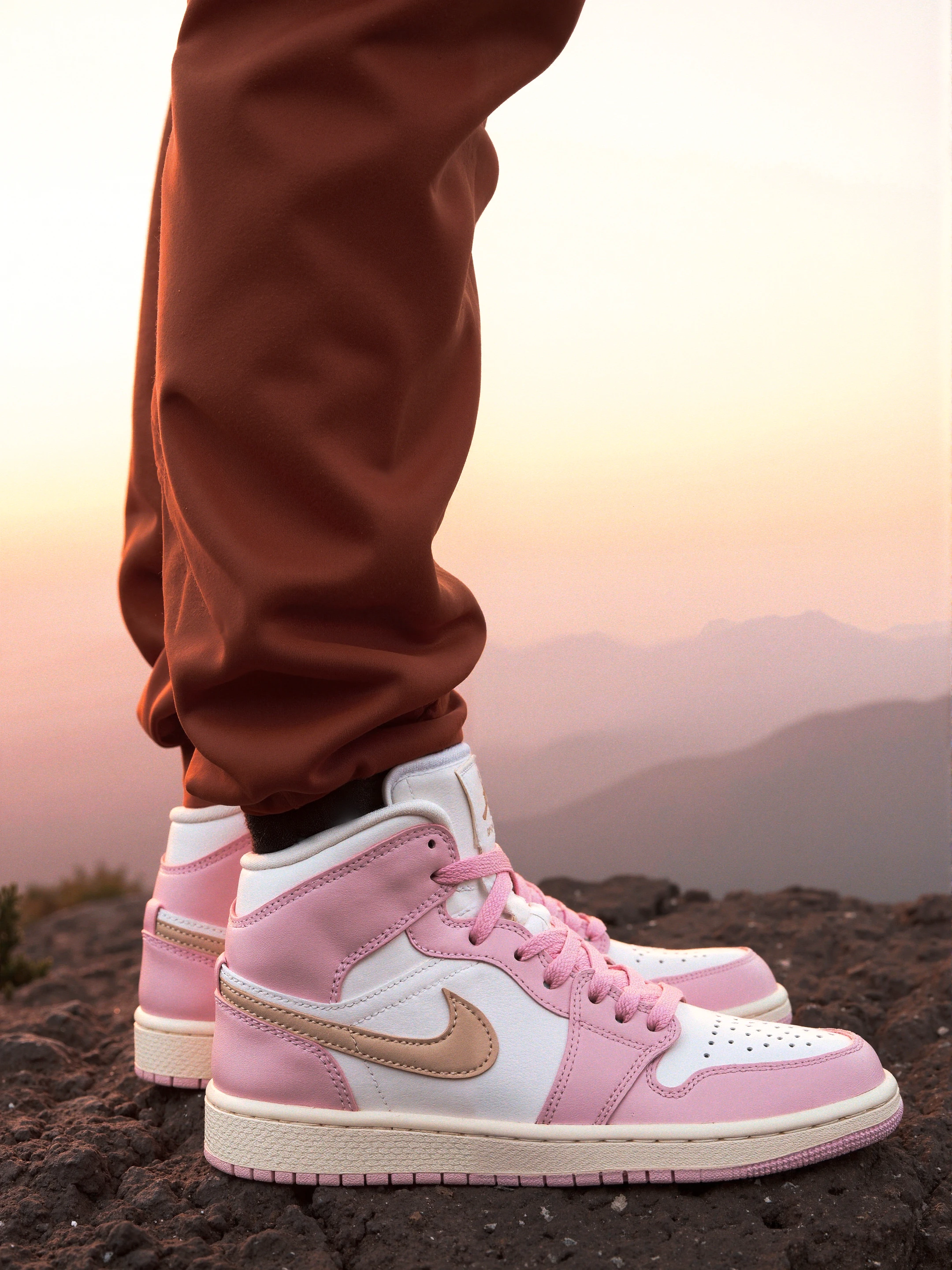 At a mountain top, golden hour, Jordan, The shoes feature a soft pink upper made of leather and synthetic materials, with a white toe box showcasing perforations for breathability. A beige swoosh logo is prominently displayed on the side. The outsole is cream-colored, complementing the overall pastel aesthetic. The laces and ankle area are also in a light pink shade, with the pants being a warm brown color. 