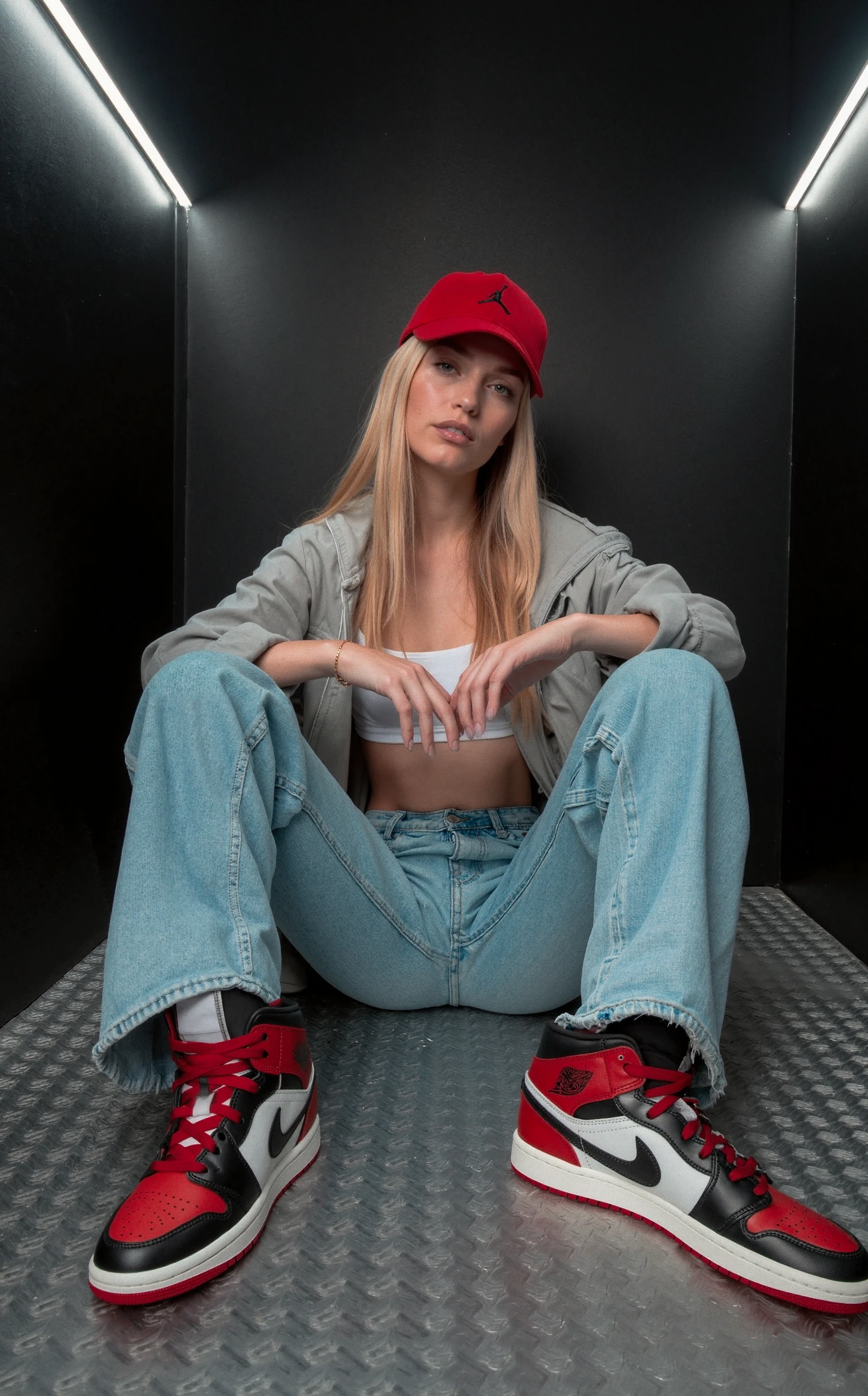 has12345.3fr,The image portrays a confident young woman seated in a casual, urban setting. She is sitting on a metallic floor, framed by sleek, dark walls, giving the scene a modern, almost industrial vibe. Her pose is relaxed but assertive; she sits with her legs spread apart, knees bent, and arms resting loosely on her thighs. Her expression is neutral, yet there is a sense of calm self-assurance in her demeanor. She wears a red backward cap over her long, straight blonde hair, adding a sporty flair to her look. The woman’s outfit consists of a light gray oversized jacket that hangs loosely off her shoulders, revealing a white sports bra underneath, paired with baggy light blue jeans. Red nike jordan sneakers, The shoe features a combination of black, red, and white colors. The upper is primarily made from leather, with black dominating the toe box and side panels. Red is present on the toe overlay, heel, and laces, while white appears on the midsection. The shoe displays a white midsole and a red outsole. The logo is a Nike swoosh in red on the side, and the Air Jordan logo is embossed in black on the outer ankle area.  stand out as the most vibrant element of her attire, matching the color of her cap and drawing attention to her feet.

The setting is minimalist, with sleek black walls that provide contrast to her outfit and add depth to the composition. The lighting is soft but direct, with light gently illuminating her face and body, casting subtle shadows that emphasize the details of her clothing and features. The color palette of the image is predominantly neutral, with gray, light blue, and black tones, while the red accents from her shoes and cap create a striking contrast, adding visual interest. The atmosphere exudes confidence and casual coolness, with a subtle rebellious undertone.