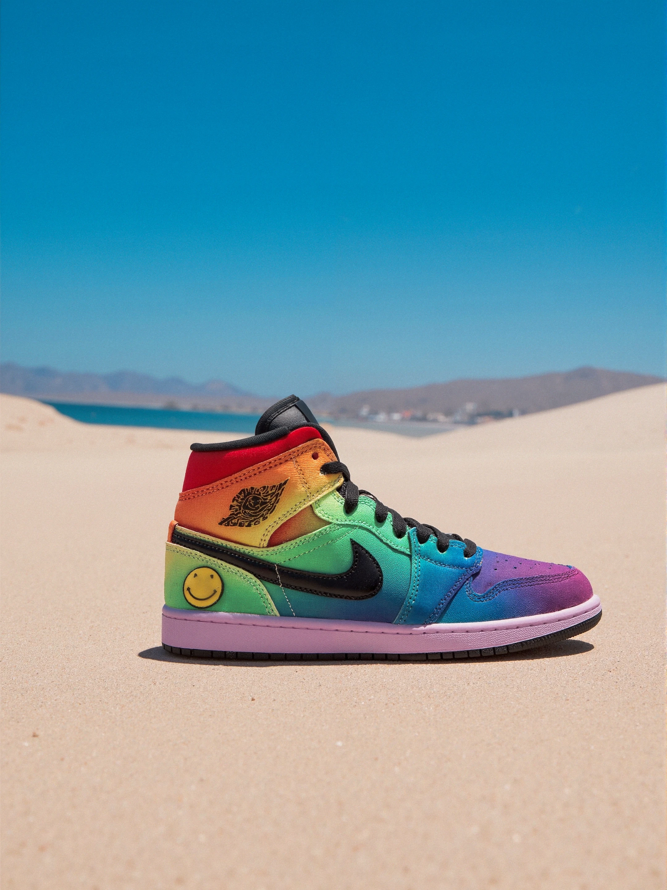 At a Beach, sand, Jordan, The shoe features a vibrant rainbow gradient with colors ranging from red, orange, yellow, green, blue, to purple. The upper is primarily made of a textile material with a smooth finish. The midsole is white and slightly cushioned, while the outsole has a pink hue. Key logos include the Nike Swoosh in black on the side and the Air Jordan logo in black on the ankle area. A playful yellow smiley face with lightning bolts is also present on the outer side of the shoe. The shoe has a high-top design with a scalloped collar. 