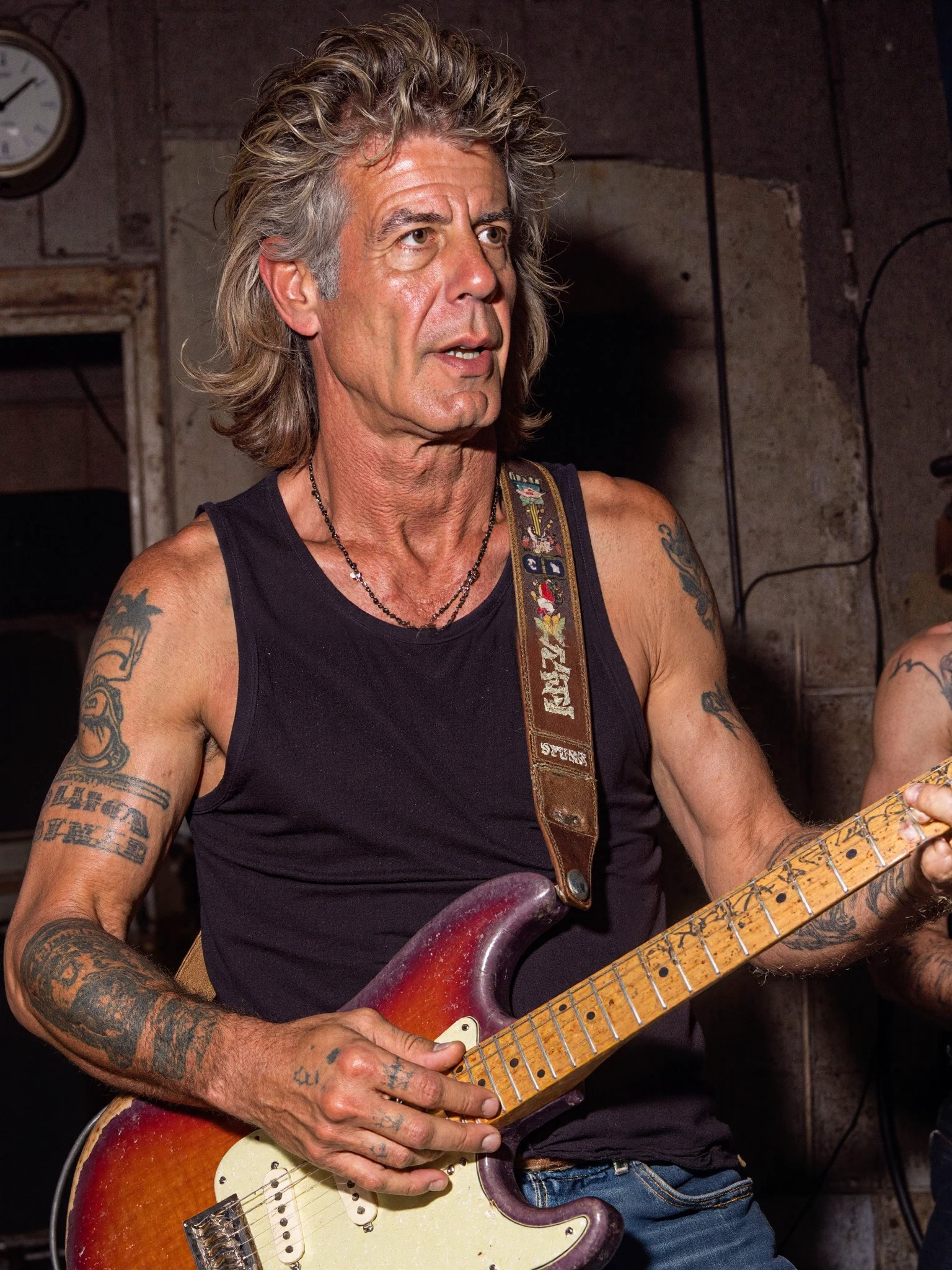 Anthony Bourdain man wearing a black tank top playing guitar in an old run down club in a punk rock band, shaggy late 1970s punk rock hairstyle, tattoos on arm, late 1970s style, punk rock, new york city club, cbgbs, cinematic, angry, 