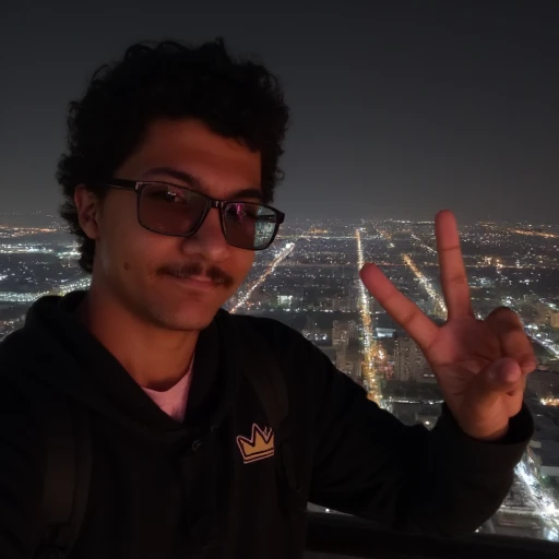 looking out at a cityscape at night. The man, curly dark hair and is wearing glasses. He is dressed in a black, dark sunglasses with rectangular frames. The man is making a peace sign with his right hand, including a crown-like design on its shoulder, and is wearing a pair of black-framed glasses. He has a mustache and is dressed in a black hoodie with a pink shirt visible underneath. The man is positioned in the foreground, revealing a lean