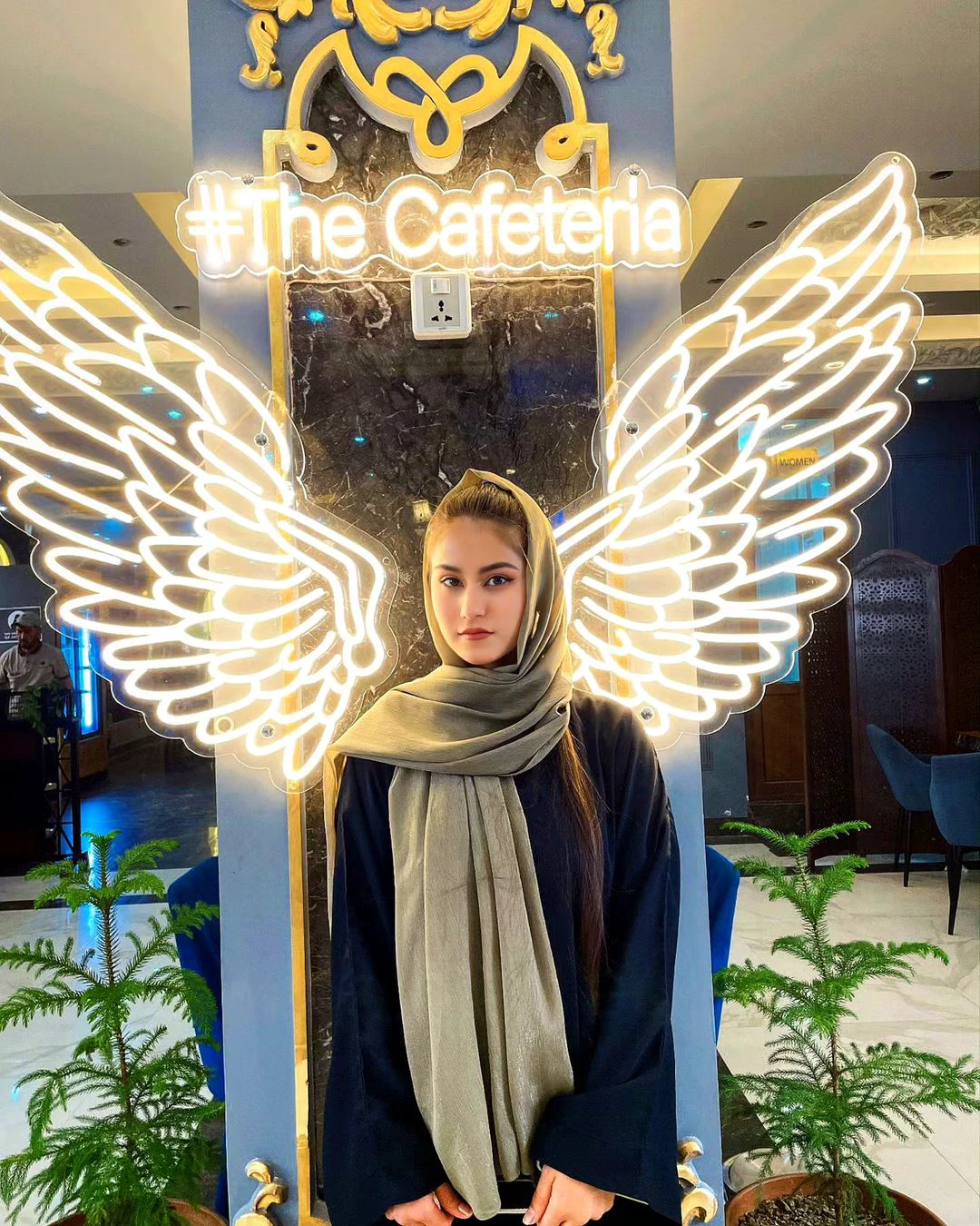 abrafluxho as a young woman with a calm expression wearing a hijab and modest clothing, standing in front of a modern cafe background. The backdrop features illuminated angel wings on a marble wall, with plants on either side of the frame. The lighting is warm and vibrant, giving the scene an ethereal and stylish atmosphere. The setting feels cozy and elegant