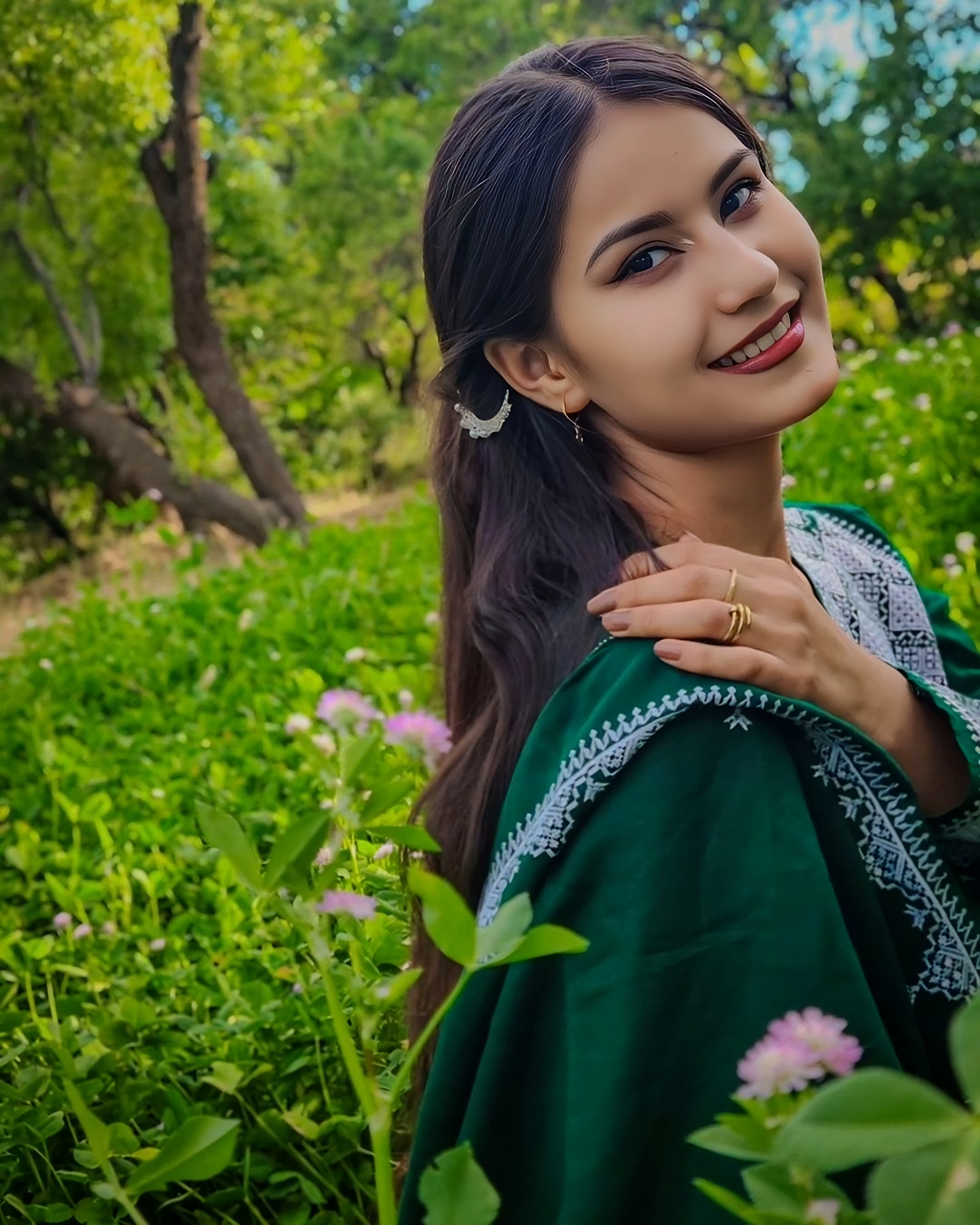 positioned in the right of the frame. The woman has a light brown complexion, This photograph captures a young woman in a lush, with a fair complexion and long, landscape. The woman, a long, rich green outfit adorned with intricate, shoulder-length dark brown hair. The woman is Hazara, petite body, smiling.