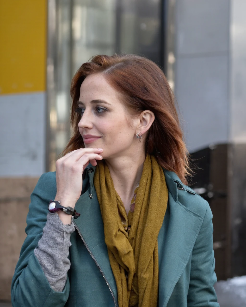 This is a photograph of eva_green with wavy, long auburn hair, captured in a candid moment. She is standing outdoors, possibly on a street corner, with a blurred urban background featuring a mix of yellow, grey, and beige colors, indicating a cityscape. Her expression is contemplative as she gazes slightly to her left, with her right hand resting on her chin. She wears a mustard yellow scarf wrapped loosely around her neck, a teal coat over a grey sweater, and a pair of purple-framed glasses tucked into her hair. Her makeup is subtle yet elegant, enhancing her natural beauty with a focus on her eyes and lips. The lighting is natural, likely in the early afternoon, with soft shadows adding depth to her features. The photograph is sharp in focus on her face and hands, while the background is softly blurred, emphasizing her as the subject.