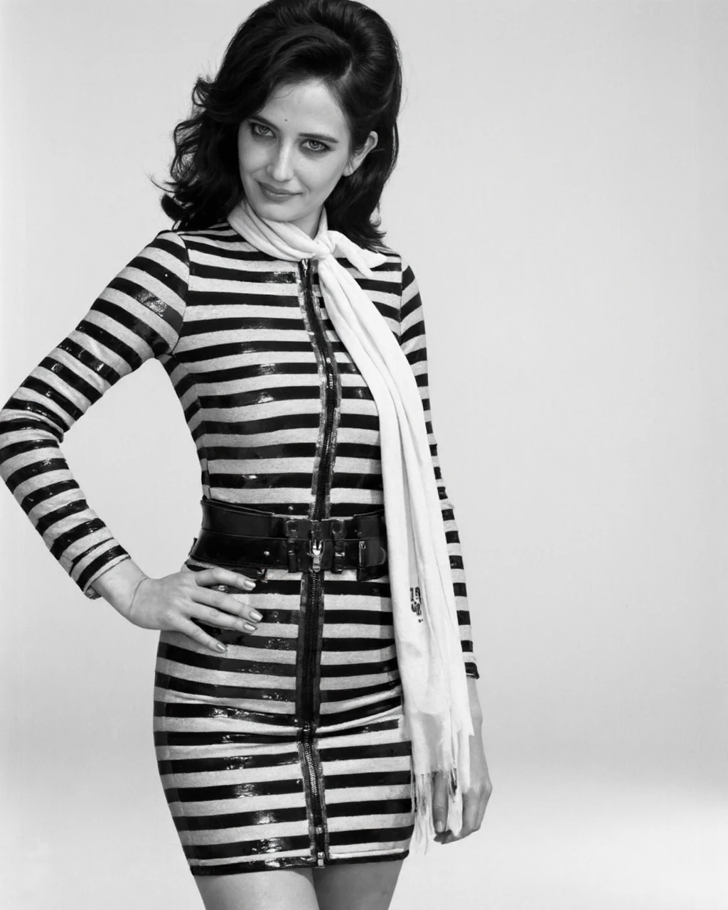 This is a black-and-white photograph featuring eva_green with long, wavy hair styled in a voluminous, retro manner, reminiscent of the 1960s. She stands in a confident, three-quarter profile, facing slightly to the right. Her outfit consists of a high-waisted, knee-length dress with horizontal stripes, cinched at the waist with a thick, black belt. The dress has a distinctive zipper running vertically down the front, adding a touch of modernity to the otherwise classic design. The sleeves of the dress are long and fitted, with a textured, possibly sequined, pattern on the upper arms. She wears a large, white scarf tied around her neck, adding a pop of contrast to the otherwise monochromatic attire. Her right hand is placed on her hip, exuding a sense of poise and self-assurance. The background is plain and devoid of any distracting elements, emphasizing the subject and her fashion-forward attire. The overall composition and styling evoke a sense of timeless elegance and individuality, characteristic of high-fashion editorials from the mid-20th century.