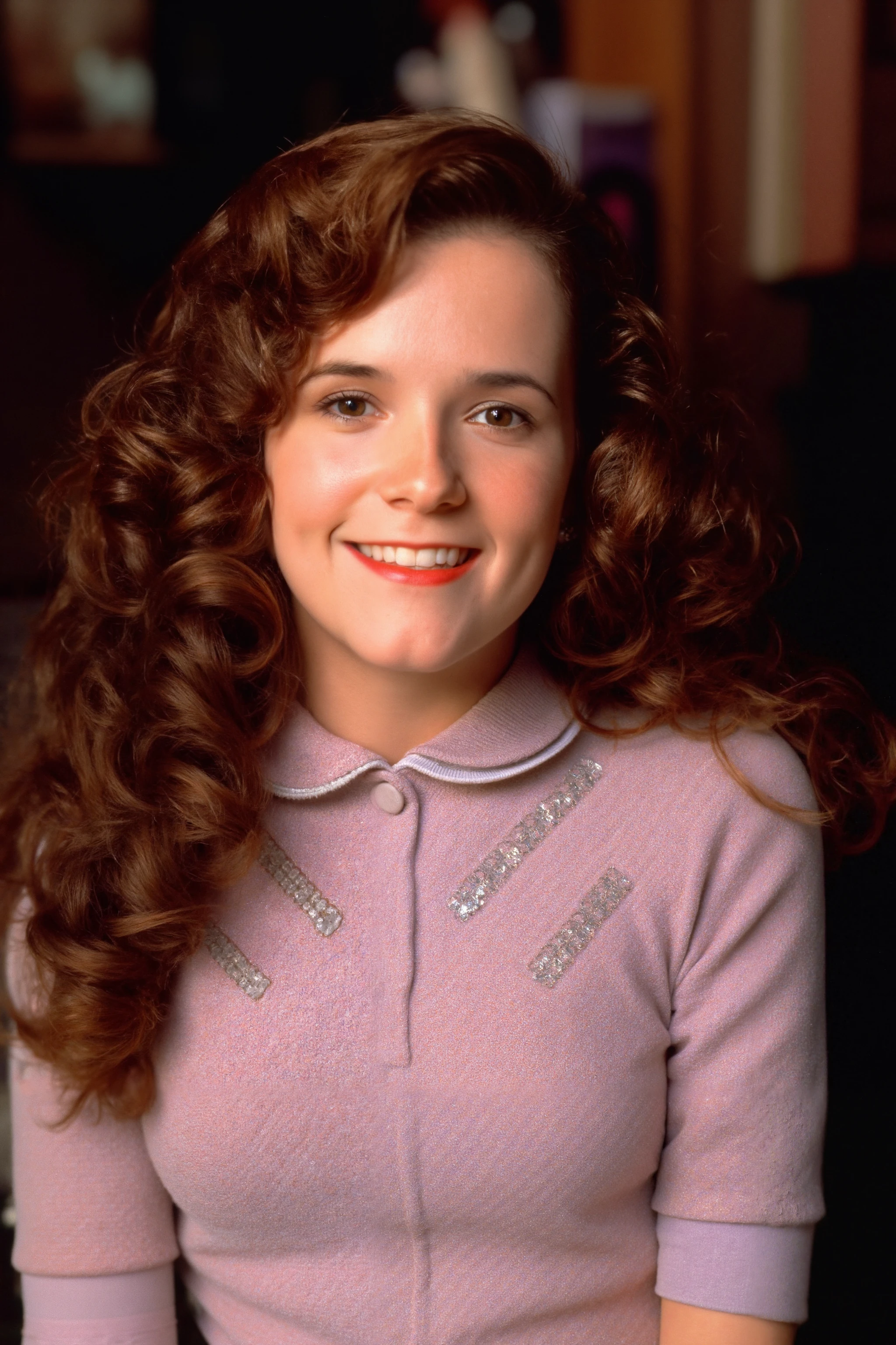 The image features a casual setting, with a girl with a fair complexion, curly brown hair, who is smiling warmly at the camera. She is dressed in a modern style outfit, high-necked dress with intricate lace details on the front.