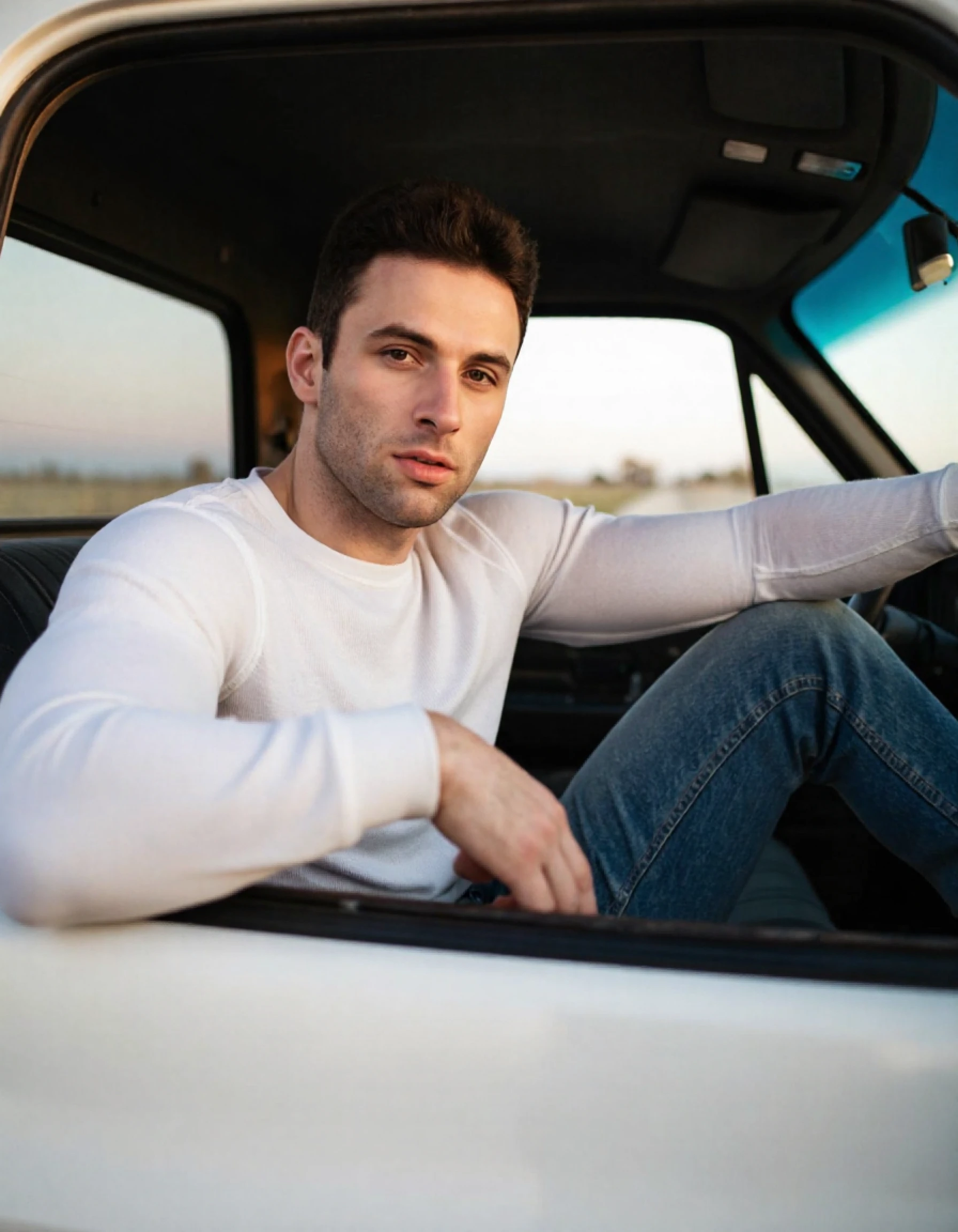 4u571n5h0w a man sitting in a car. he is handsome. he is muscular. he is wearing a white henley. he is wearing blue jeans. he is looking at you. he is modeling in the car. he has his arm on the top of the seat. it is dusk. the car is an older truck.