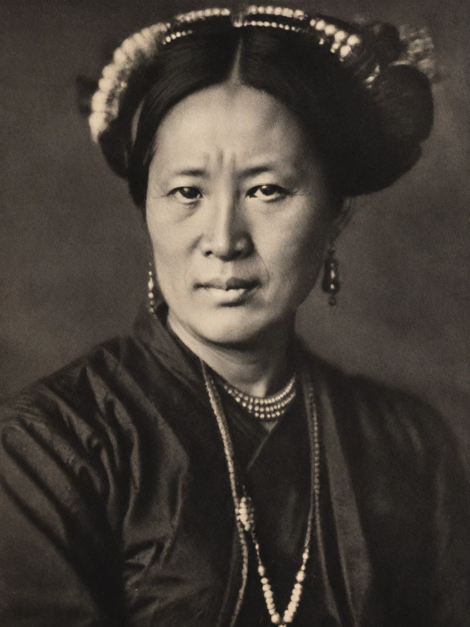 pictorialist portrait photo in the style of alvin-langdon-coburn, A Mosuo woman from China, in her 50s, with a calm, serene expression, dressed in a traditional costume with a headpiece and heavy silver jewelry, monochrome
