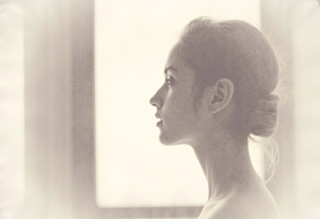 an aesthetic, minimalist depiction of a female profile in side view. The focus is on the soft contours and calm, monochromatic tones. The scene feels mystical and dreamy, almost as if viewed through a delicate mist, with gentle light accentuating the silhouette. The background is diffuse and creamy white, drawing attention to the elegant simplicity of the figure.