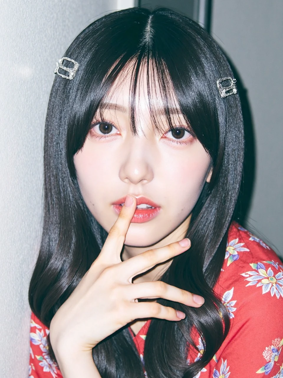 The image is a young woman named konishi with long dark hair. She is wearing a red blouse with a floral pattern and has two silver hair clips on her head. Her hair is styled in loose waves and thin fingers. Her fingers are very slender and she touches her lips with them. She has a serious expression on her face and is looking directly at the camera. The background is gray white walls, and the environment is dim. The dim lighting adds a touch of mystery to the characters.