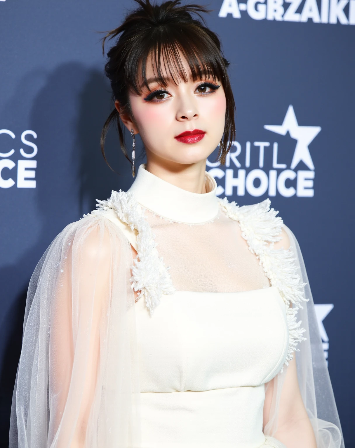 This is a high-resolution photograph of a young woman standing against a backdrop with a blue-gray color scheme. The backdrop features the words "CRITICS CHOICE" and a row of stars, indicating a red carpet event. The woman has fair skin and dark brown hair styled in a loose, elegant updo with a few loose strands framing her face. Her eyes are accentuated with dark, smoky eye makeup, and she wears deep red lipstick that complements her dark eyelashes. She is dressed in a sheer, white lace top adorned with intricate patterns and small, sparkling embellishments. The lace top has a high, ruffled collar that adds a touch of vintage elegance to her outfit. Her earrings are small, round, and silver. The background is slightly out of focus, emphasizing the subject. The overall atmosphere of the image is sophisticated and glamorous, typical of a high-profile event.