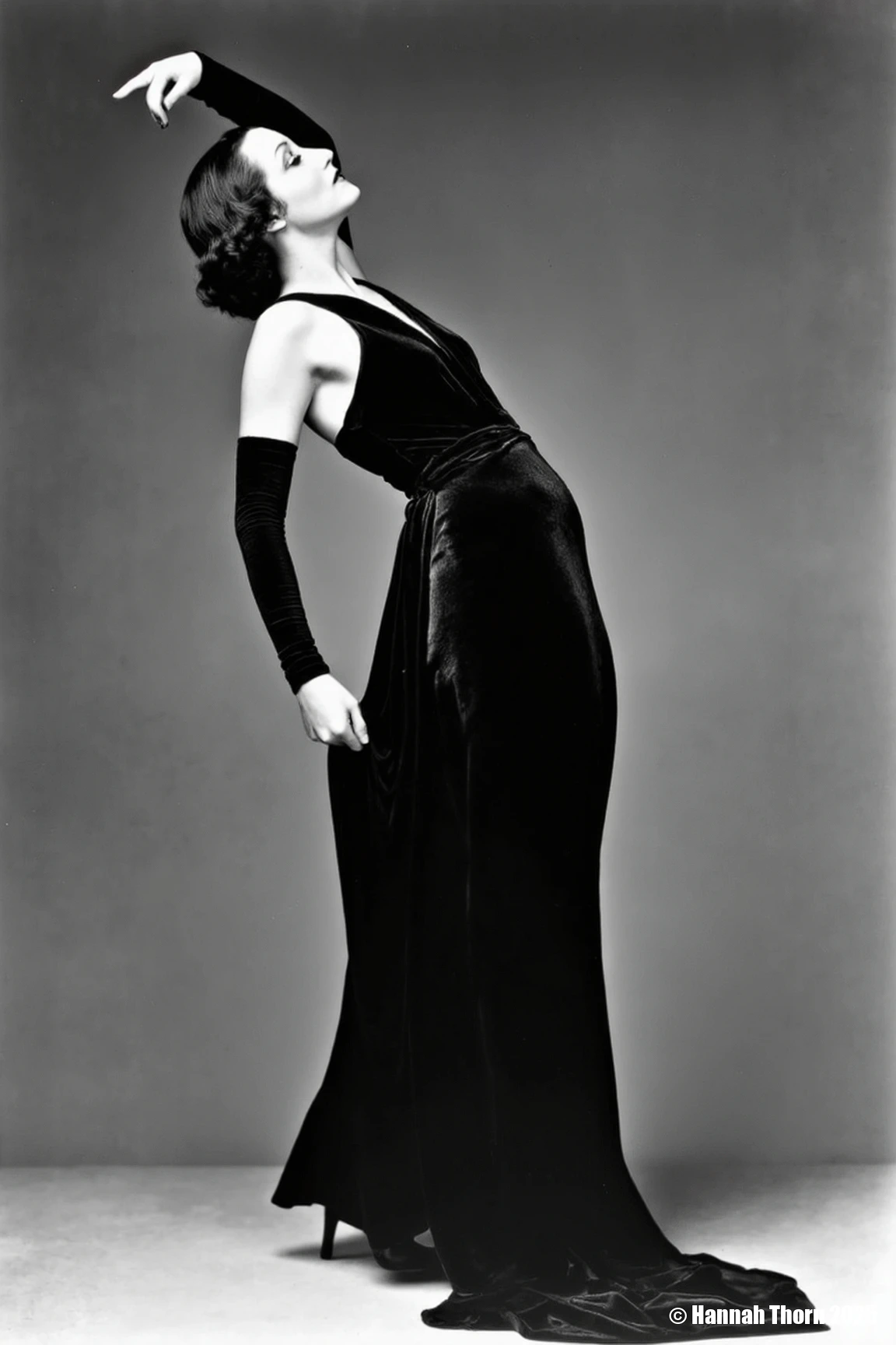 This black-and-white photograph captures a sideview of a woman in a striking, dramtic pose of elegant, stylized sensuality. The woman's body is arched slightly backward, her head tilted upward and to the side. One arm is raised above her head, her hand gently touching her hair; the other arm hangs down, but is bent at the elbow and she is using her fingers to hold the fabric of her dress at her waist. Her expression is serene and confident, with a hint of mystery. The overall effect is a graceful, almost languid pose that conveys both confidence and allure, suggestive of a classic Hollywood starlet. The pose is carefully crafted to showcase the figure and the gown. The gown is a dark-colored, possibly black, velvet or similar fabric. It's a sleeveless design with a very low, possibly plunging, neckline that reveals much of the back and shoulders. The style is form-fitting, hugging the body's curves and creating a sleek silhouette. The dress features a keyhole or cutout detail at the neckline and long sleeves that extend into what look like fitted gloves or long opera-length gloves. The fabric appears to have a subtle sheen or texture, suggesting a luxurious material. The overall design is suggestive of 1920s or 1930s Hollywood glamour., detailifier<lora:_Flux\detailifier_flux-000006.safetensors:0.0:0.0>