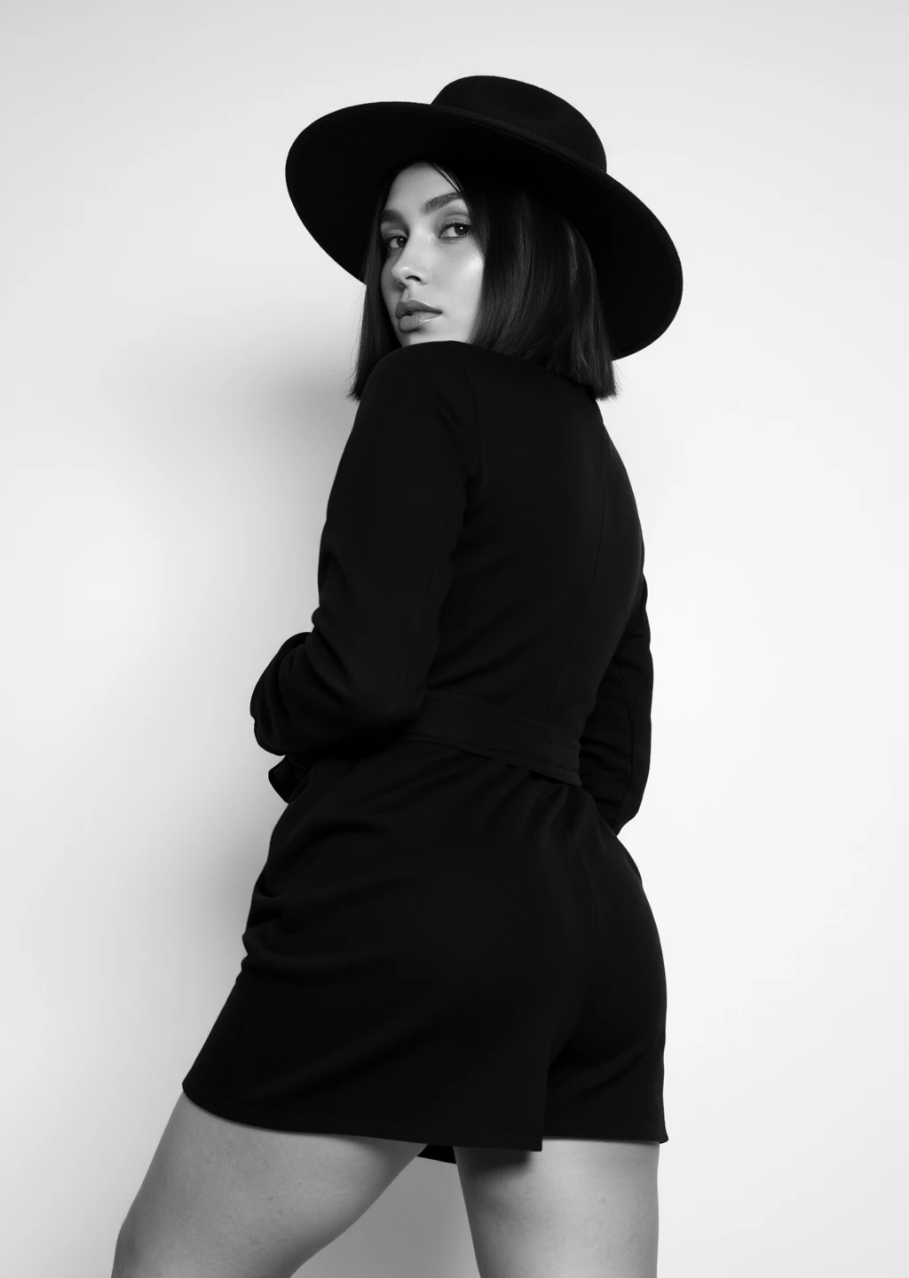 fpose-photography, This is a black-and-white photograph featuring a confident woman from side, in a stylish pose with slightly leaning backward . She is looking away from the camera .  She has a sleek, shoulder-length bob haircut and wears a large, black hat. Her outfit includes a fitted black blazer and a short skirt . background is a plain, light wall, casting a soft shadow of the woman. The image's monochromatic palette 