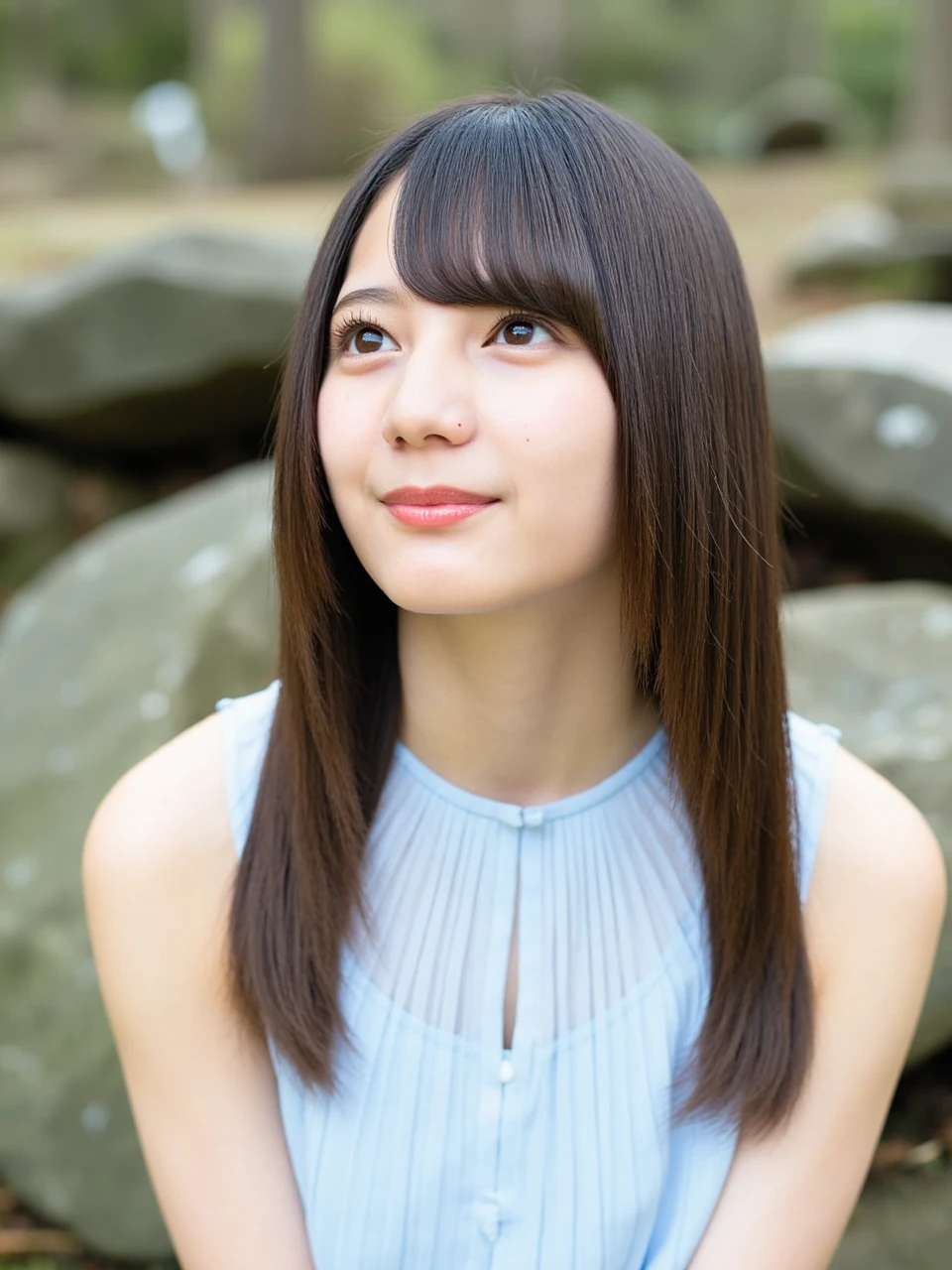 The image is a portrait of a young woman named kosakana with long hair and bangs. She is wearing a light blue dress with a halter neckline. The dress is made of a sheer fabric with a pleated design. The woman is looking up towards the sky with a dreamy expression on her face. The background is blurred, but it appears to be an outdoor setting with rocks and greenery. The lighting is soft and natural, creating a peaceful and serene atmosphere.