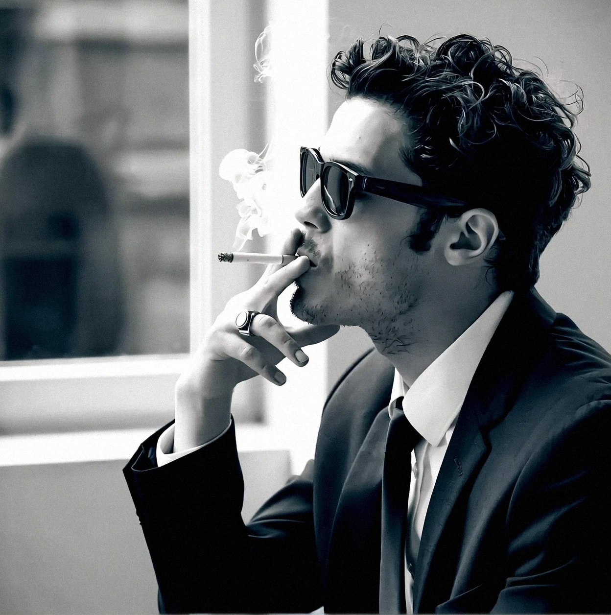 A black and white photo shoot from a profile camera angle about a man in a formal suit smoking a cigarette while wearing sunglasses. a young man with curly hair, wearing a black suit and a white shirt with a black tie, sitting in the middle of the image, with his hand resting on his chin and a cigarette in his mouth. he appears to be in his late twenties, with a serious expression and a slight stubble on his face. smoke is coming out of his mouth, indicating that he is smoking. he is wearing sunglasses and has a ring on his finger. the background is blurred, but it seems to be an indoor setting with a window, suggesting that the photo was taken indoors. the image has a monochromatic tone, and the focus is on the man's contemplative expression and the smoke.
1boy, solo, looking at viewer, short hair, black hair, suit, tie, collared shirt, smoking, sunglasses, suit jacket, tie necktie, ring, cigarette, smoking cigarette, ring on finger
camera_angle: profile, art_style: photo realistic, location: indoor, background: blurred interior setting with windows, suggesting a formal setting, text: NA, distance_to_camera: upper body, clothing: black suit, white shirt, image_composition: middle, pants: n/a, accessory: sunglasses, shoes: N/A, action: smoking, facing_direction: facing viewer, eye_direction : looking to the side, facial_expression: contemplative, ear: slim, gender: 1man, hair_color: curly hair;black hair, hair _style: short hair;short hair, race: NA;NA, body: slim