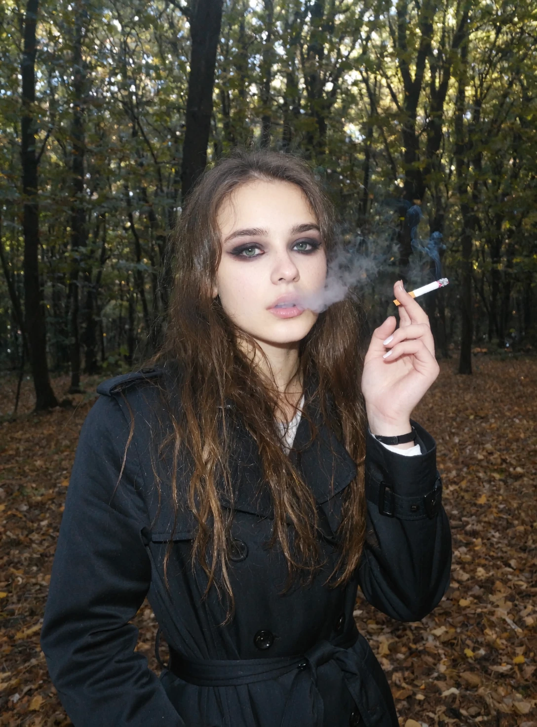 l3n0v0. underexposed outdoor scene, teenager girl, casual style, wearing dark trench coat, long wavy brown hair cascading over her shoulders, looking direct at the viewer, gothic heavy makeup, outdoors in the dark forest, autumn, smoking cigarette with smoke, showing middle finger not point finger, sunlight filtering through the trees. amateur quality, low resolution, candid style