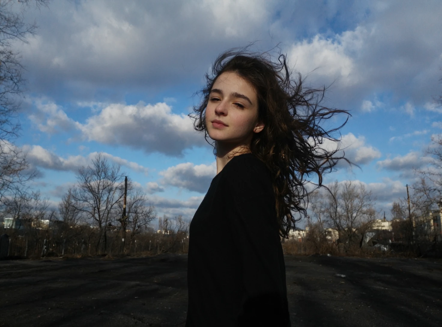 l3n0v0. portrait of teenager girl, flowing tousled hair, from side view angle, looking at the viewer, dreamy expression, closed eyes, bare winter trees, dramatic cloudy sky, bright sunburst through clouds, deep blue sky patches, industrial urban setting, dark shadowed foreground, late afternoon lighting, silhouetted landscape, underexposed ground elements, natural dramatic lighting, high contrast image. amateur quality, candid style
