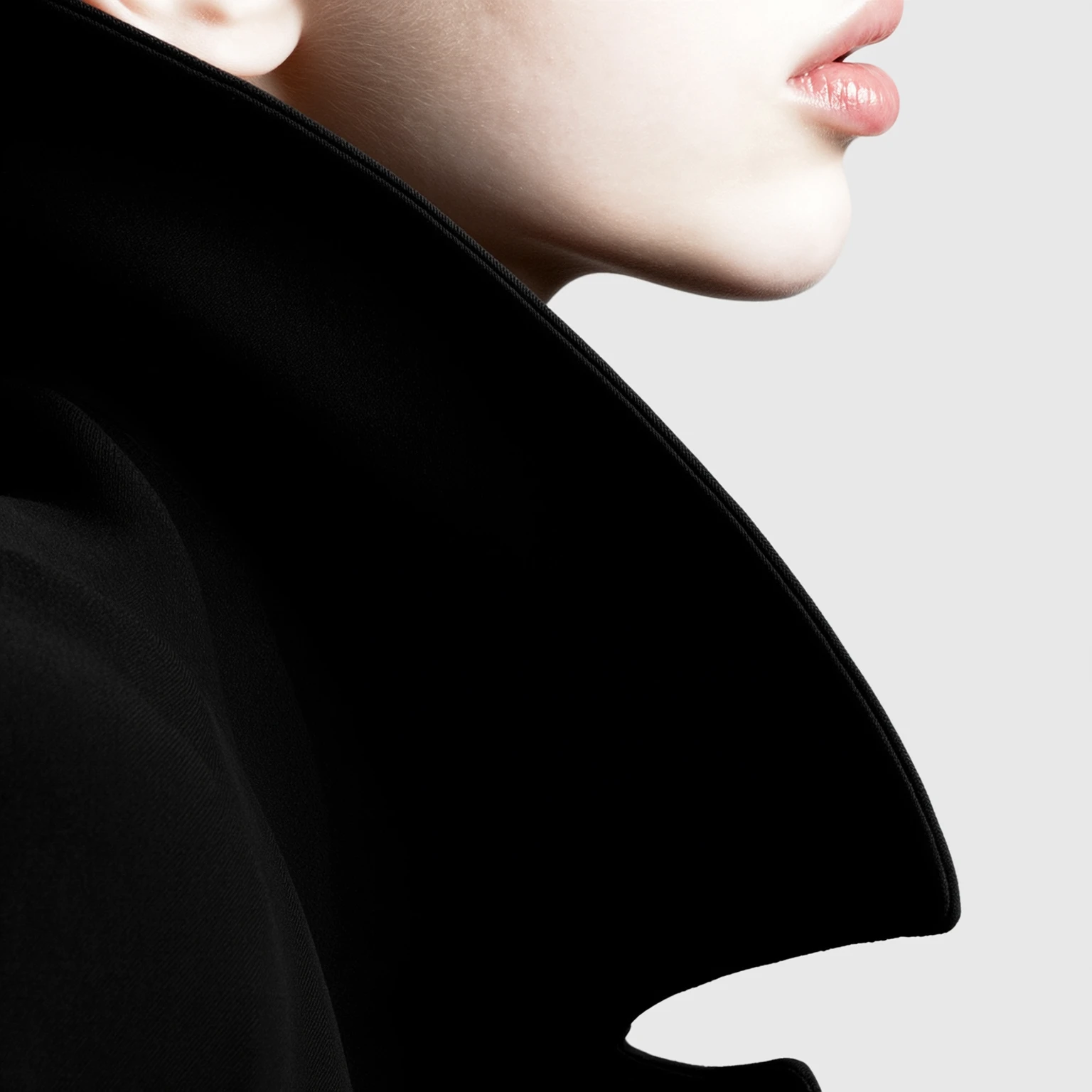 写实风格的艺术人像特写，浅色皮肤女性侧脸局部，画面聚焦在嘴唇、下颌、颈部和部分耳朵，双唇自然闭合，皮肤细腻光滑，头发浅色，佩戴一件高挺的黑色大衣，衣领向上翻起形成锐利的几何线条，背景为干净的浅灰色，影棚级戏剧化打光，黑白强烈对比，光影在皮肤与衣物之间形成鲜明分界与细腻层次，构图极简、线条流畅优雅，画面具有现代时尚与抽象艺术摄影的高级质感，超高清分辨率