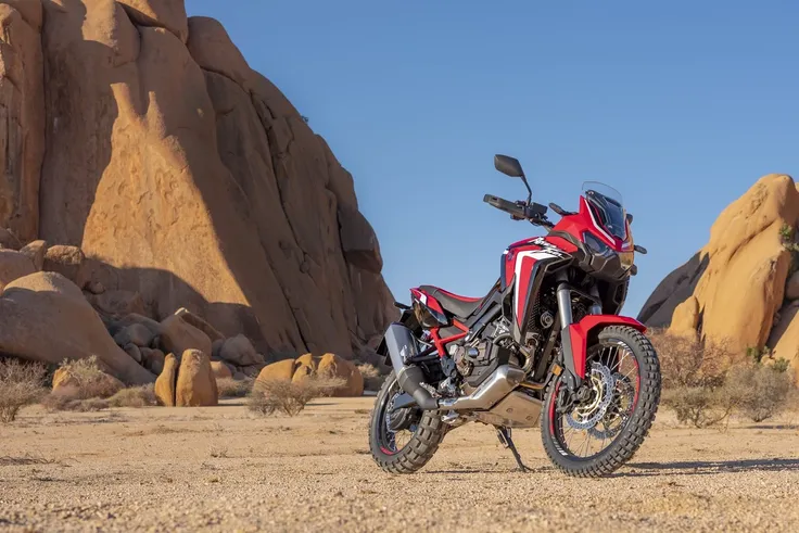 Standing alone arafed adventure motorcycle with desert mountains background, big windscreen, mud dirt tyres, one exhaust, honda, dakar motorbike, red fairing, skid plate