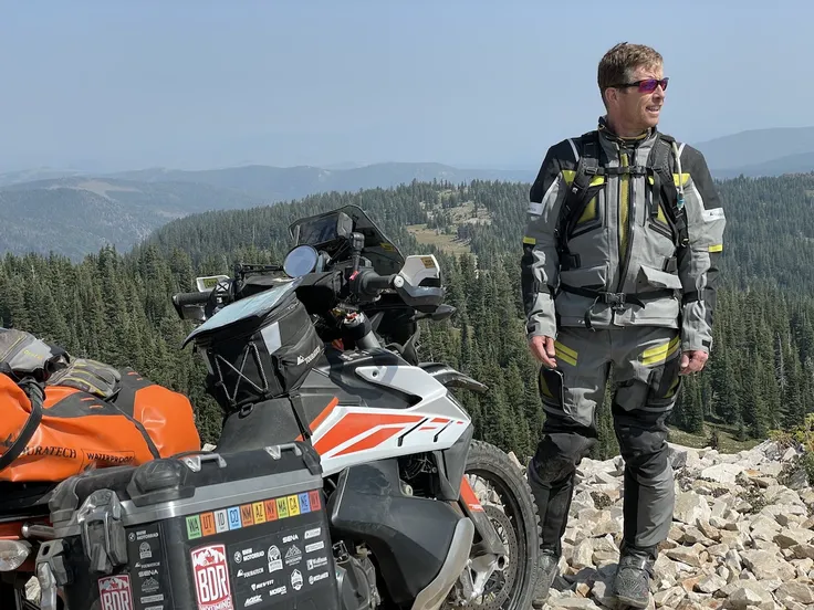 arafed rider with adventure gear standing next to a motorcycle, forest and mountains background, no helmet, wearing boots