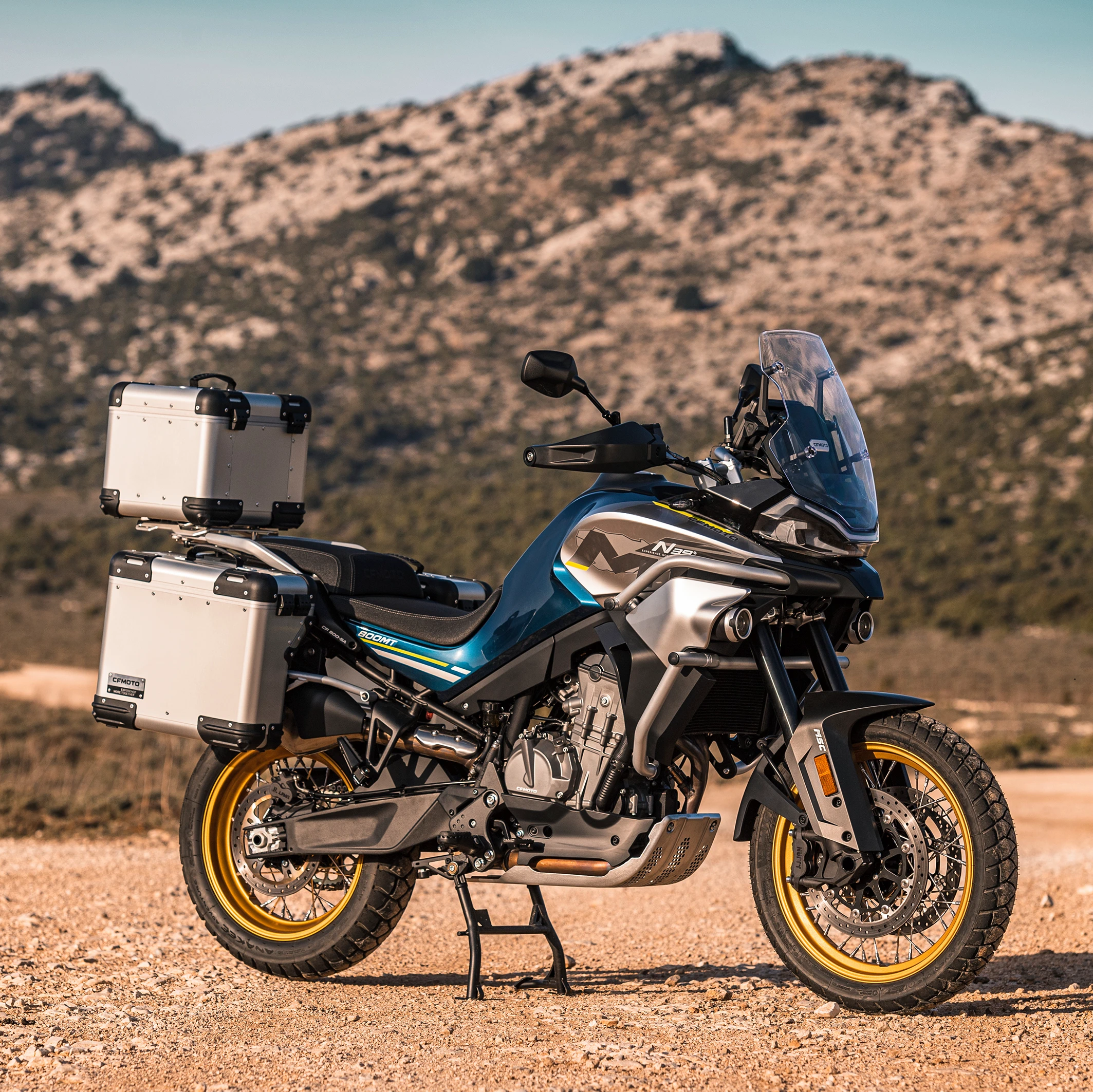 Standing alone arafed adventure motorcycle with mountains background, big windscreen, mud dirt tyres, one exhaust, rear luggage, triumph, blue and silver fairing, skid plate