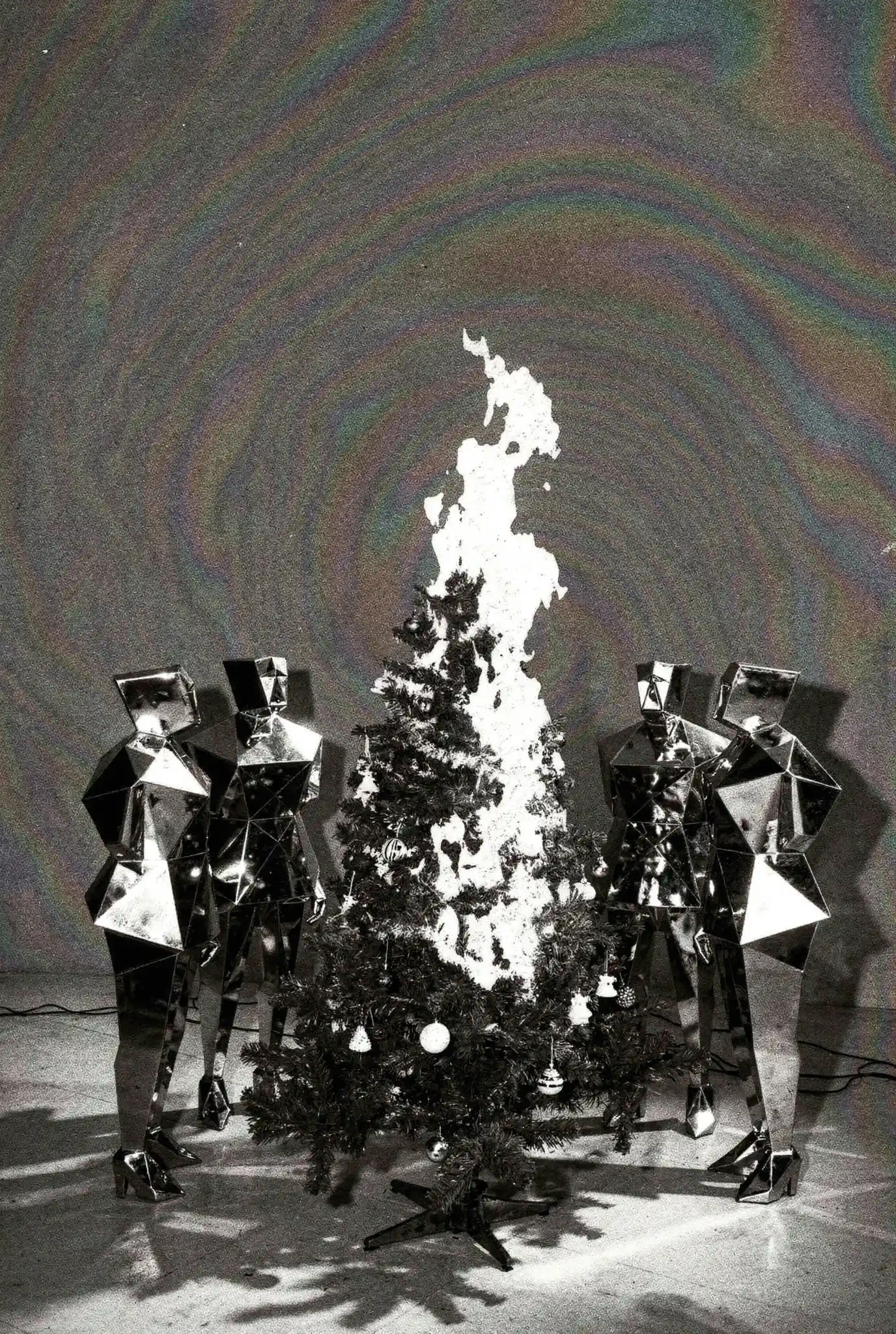 Two figures in geometric mirror costumes surrounding burning Christmas tree with holographic rainbow backdrop