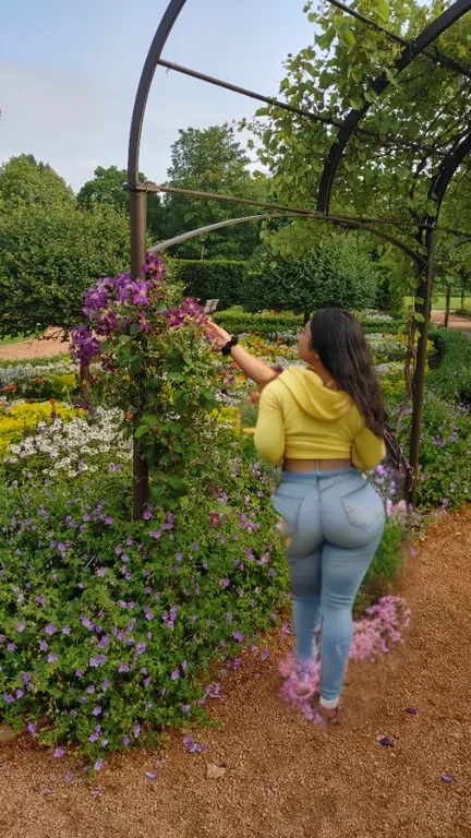 woman in yellow shirt standing in a garden with flowers, standing in a botanical garden, standing in flower field, standing in a flower field, picking up a flower, parks and gardens, picking flowers, girl standing in a flower field, ancient garden behind h...