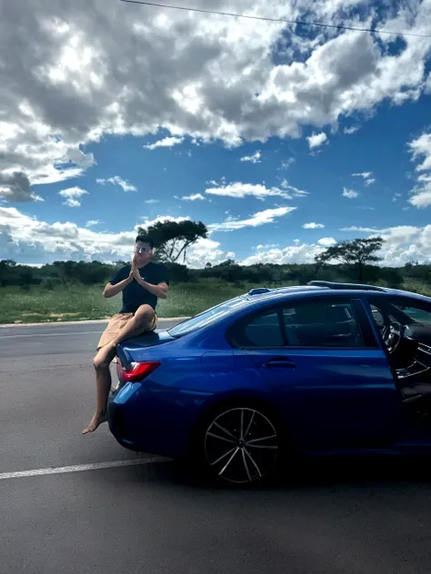 a boy smiling, on top of a car, with prescription glasses, on a road, with a very blue sky, but with clouds and a radiant sun