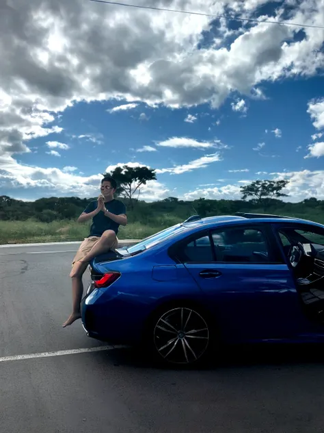 a boy smiling, on top of a car, with prescription glasses, on a road, with a very blue sky, but with clouds and a radiant sun