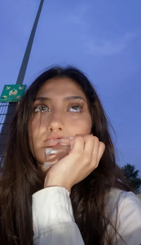 A girl with brown eyes and dark hair, with a Middle Eastern look, is smoking a cigarette by the pool on a summer day.