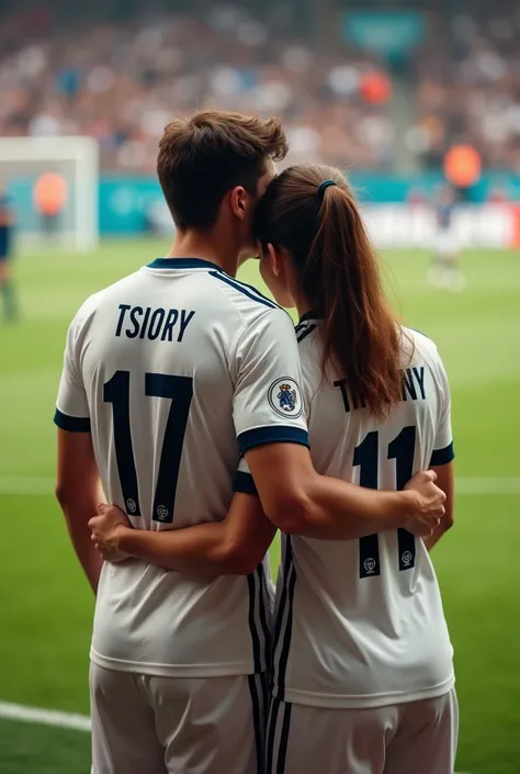 Un jeunne homme porte le maillot de real madrid anomme "TSIORY"au dos ,numero 17 regarde un match au terrain de foot,et une fille porte le maillot de real madrid anomme "TIFFANY" au dos ,numero 11 lembrasse