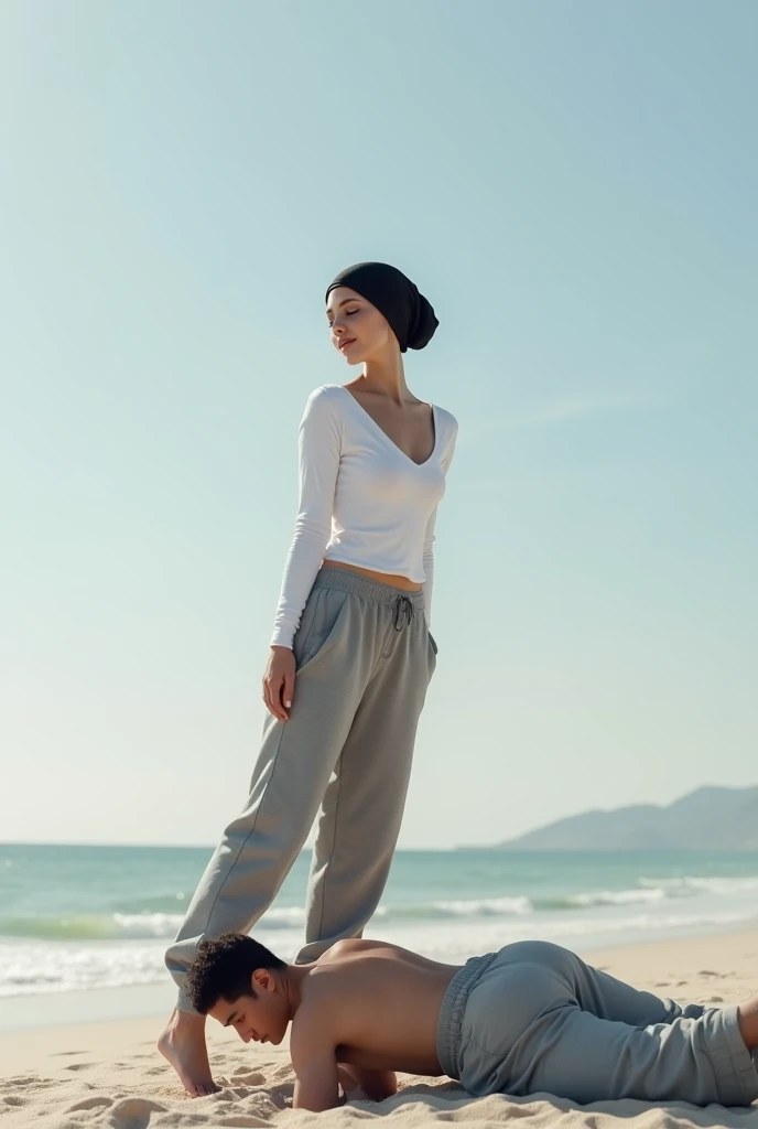 Black headscarf on the beach, grey baggy sweatpants, white long sleeve t shirt, barefoot, sexy and skinny woman. Man lying on his stomach in front of woman. Woman puts her foot on the front of man&#39;s sweatpants. 