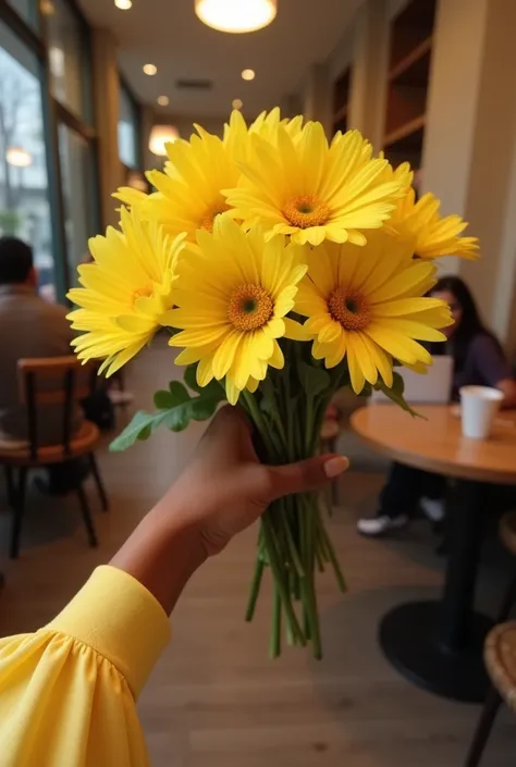 point of view of a female brown hand, with long sleeves holding a bouquet of yellow flowers with a cafe background 