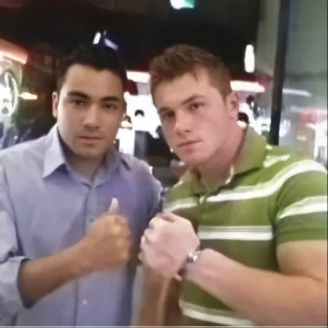 Two young men are standing side by side in a friendly yet intense pose. The man on the left, with medium-light skin, wears a light blue or gray button-up shirt, his dark short hair neatly styled. The man on the right, with fair skin, sports a green and whi...