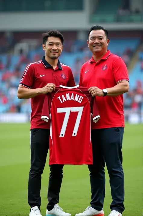 Photo of Shin tae young, the Indonesian football coach, next to a fat Malay man with neatly trimmed hair wearing a RED polo shirt, they are holding a football shirt that says "TATANG 77". He was standing on the grass field. The background contains a stadiu...