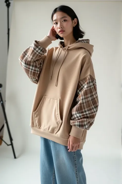 A girl is taking a photo in a studio as a model. She is wearing an oversized hoodie in cappuccino color. The hoodie has unique sleeves made of different plaid fabric with small shirt cuffs at the bottom. The plaid is white underneath, with beige, brown, an...