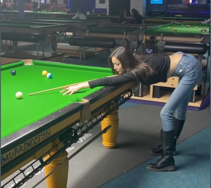 This photo was taken in a modern, well lit billiard hall with high resolution. The central figure is a young woman with long and straight black hair, leaning against a green felt pool table, holding a billiard club in her hand, bending down and staring int...