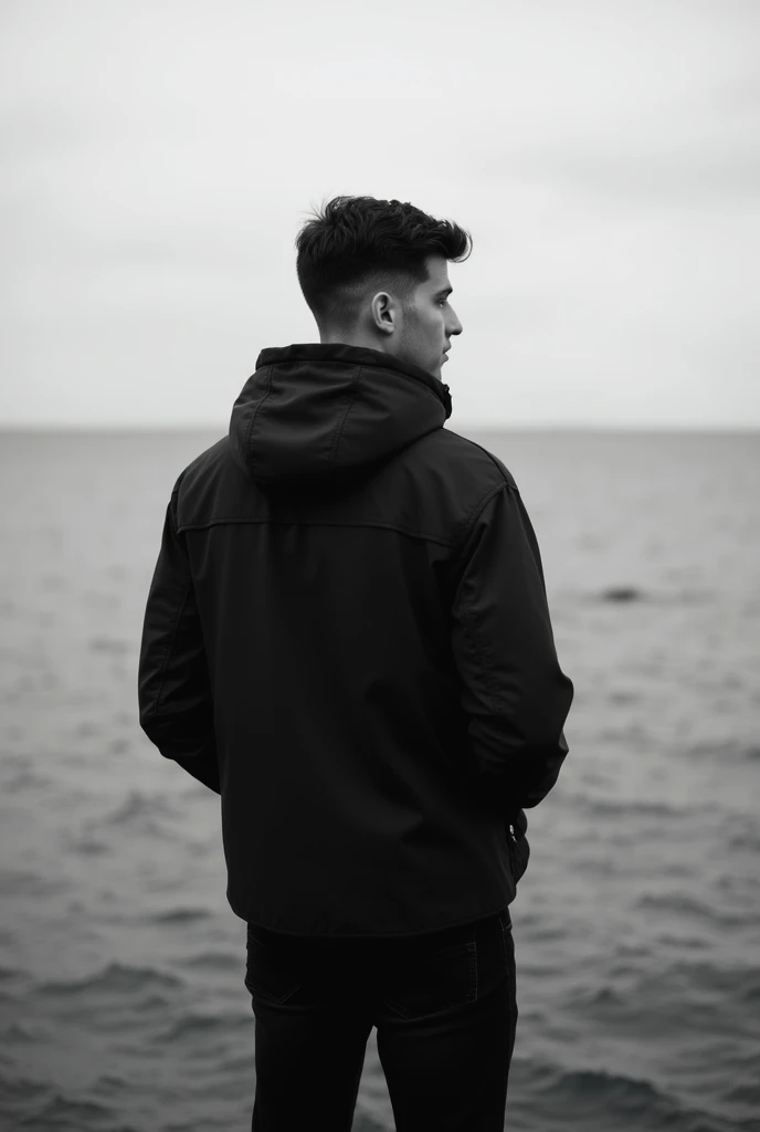 A black and white Instagram profile picture of a young man looking at the sea wearing and 
The jacket has a hat on 
 A black jacket and pants and the photo is from the back 