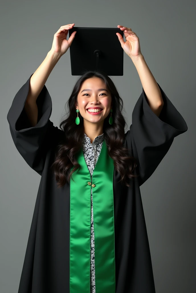 A young woman dressed in a black graduation gown with green accents, holding her graduation cap above her head with a joyful expression. She has long, dark hair styled in loose waves, and is wearing statement green earrings that match the gowns sash and li...