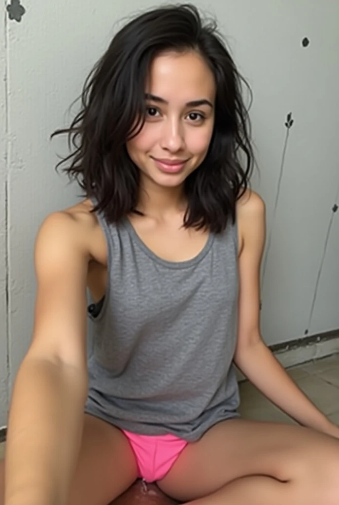 A selfie of a young, dark-haired woman, wearing an oversized gray tank top and pink shorts, sitting on the floor. The background is a light gray wall with a simple black flower pattern. The image has a slightly grainy quality, suggesting a cell phone camer...