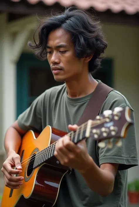 a 30 year old Indonesian man with curly shoulder-length hair wearing a modern t-shirt is playing his guitar in front of his house